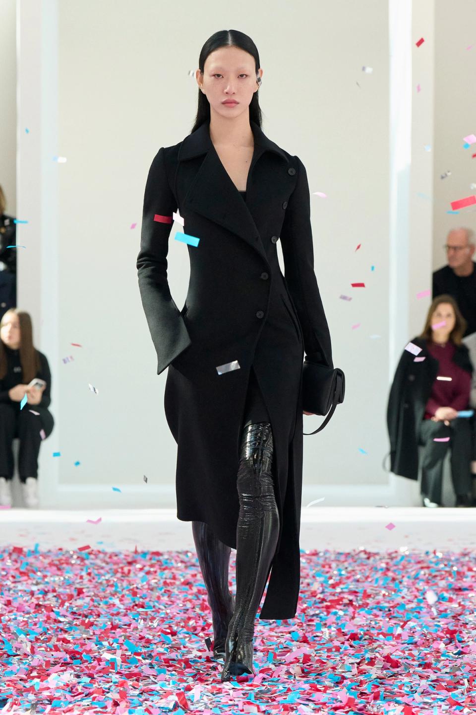 Elegant model wearing a black Courrèges coat on runway with colorful confetti, for ecommerce.