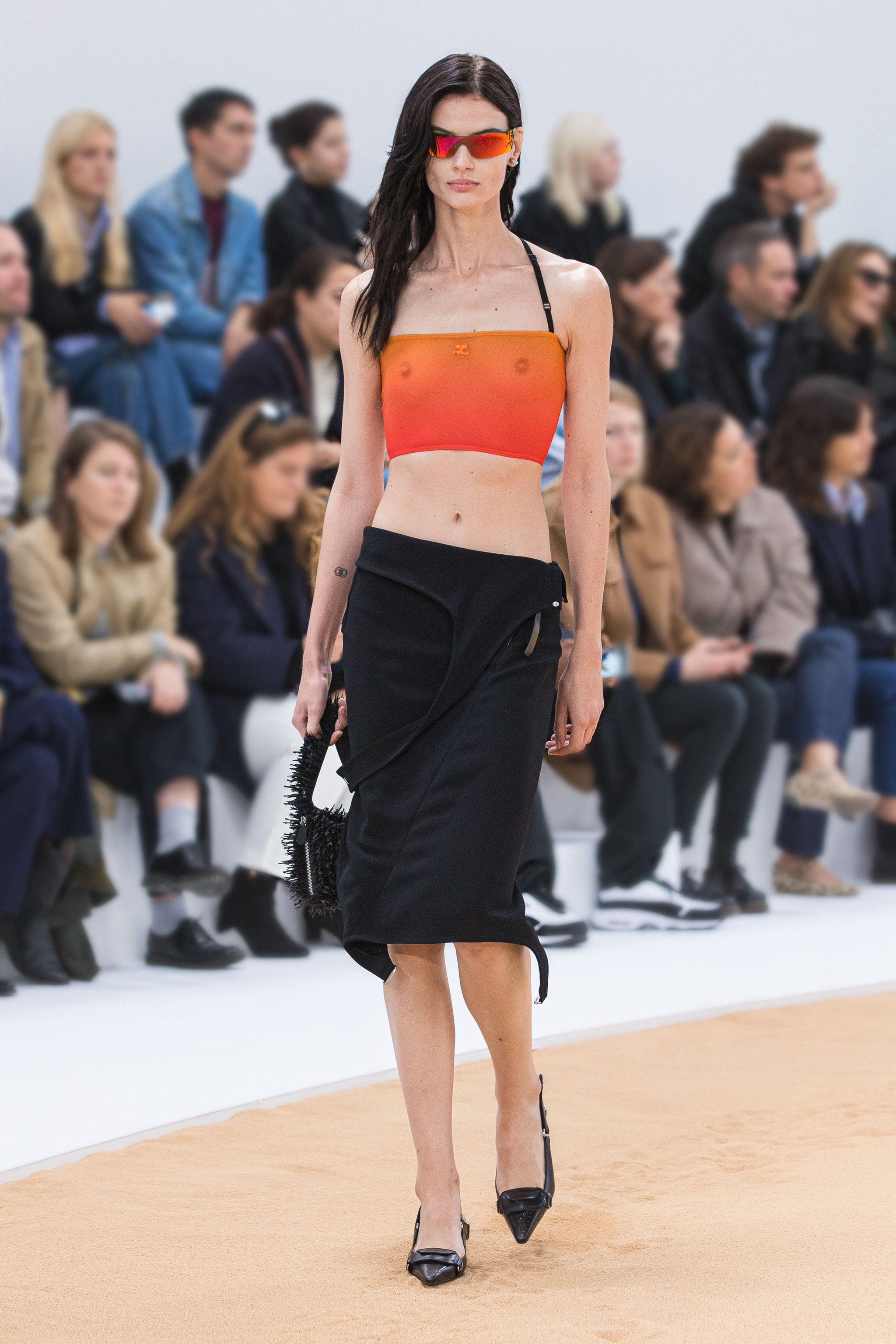 Elegant model on runway wearing Courrèges fashion, black skirt, orange top, and stylish accessories.