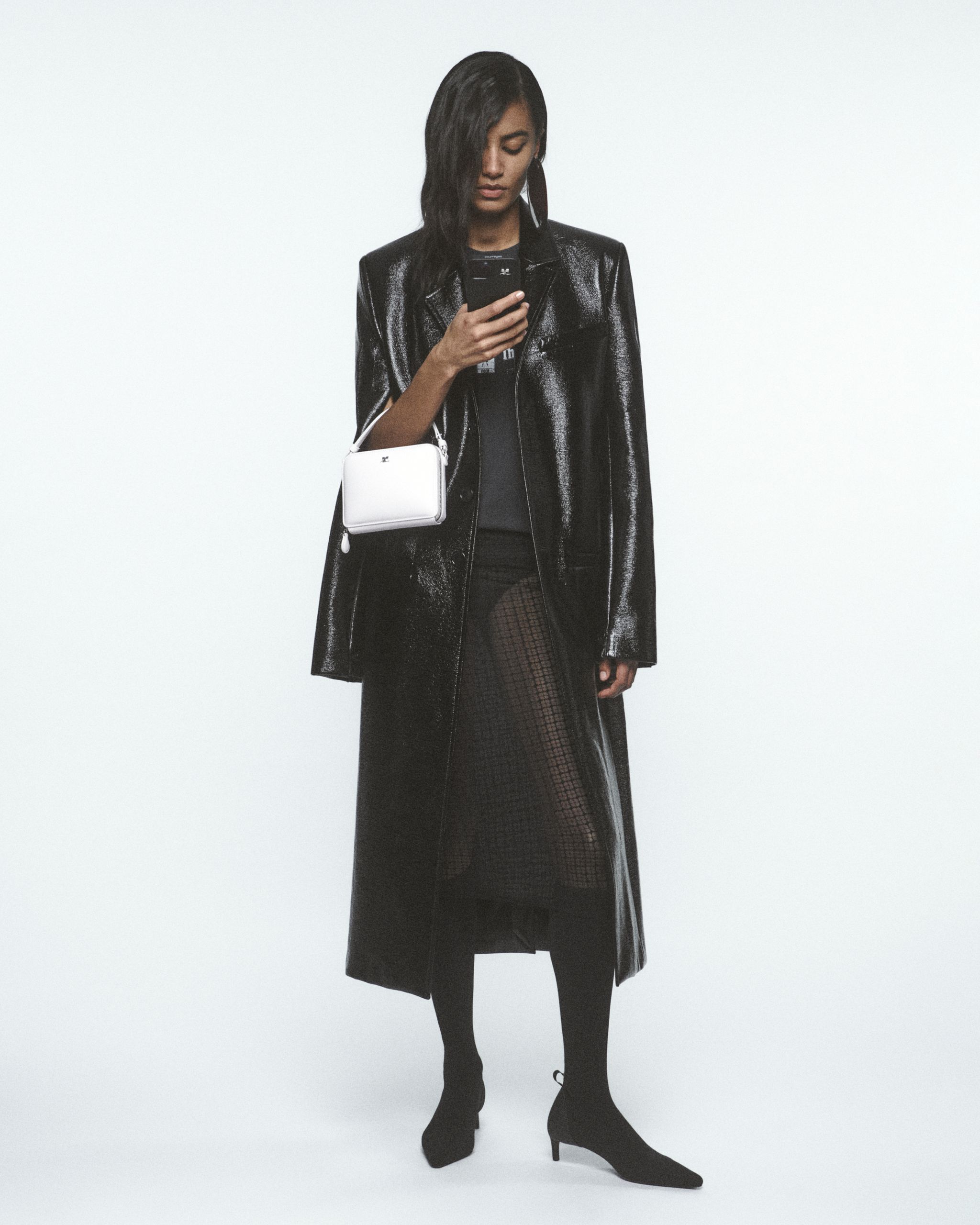 Femme élégante en manteau en cuir noir, collants en filet, portant un sac Courrèges, mode pour ecommerce.