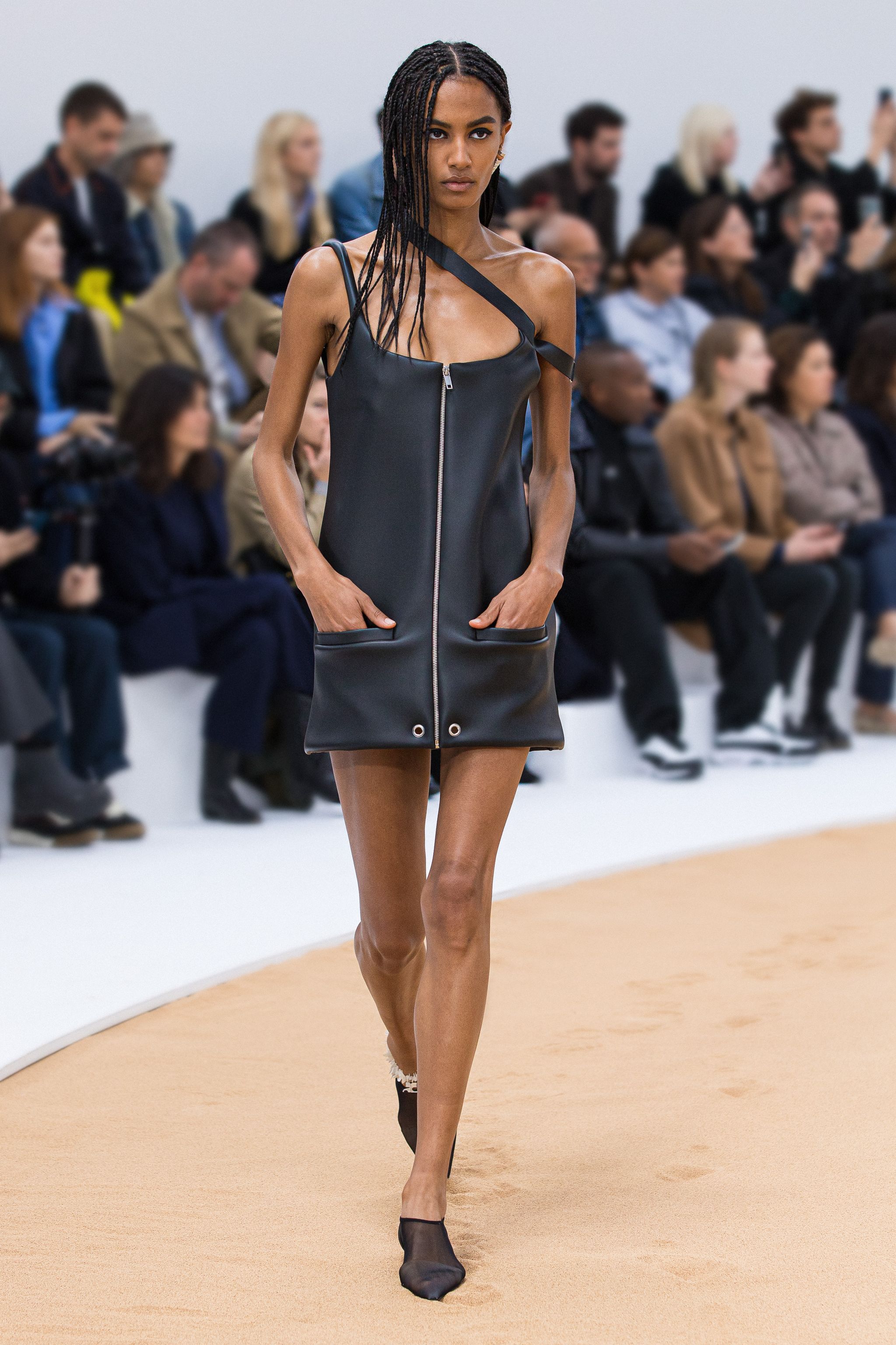 Fashion model walking on runway wearing Courrèges black leather dress with zipper, audience in background.