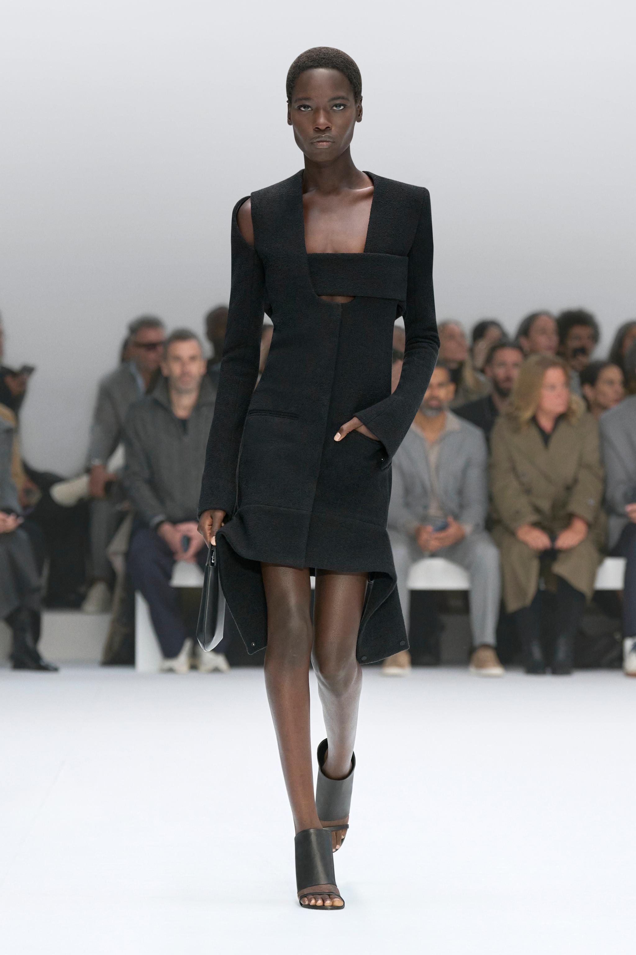 Elegant black dress by Courrèges showcased on a runway with an audience in the background.