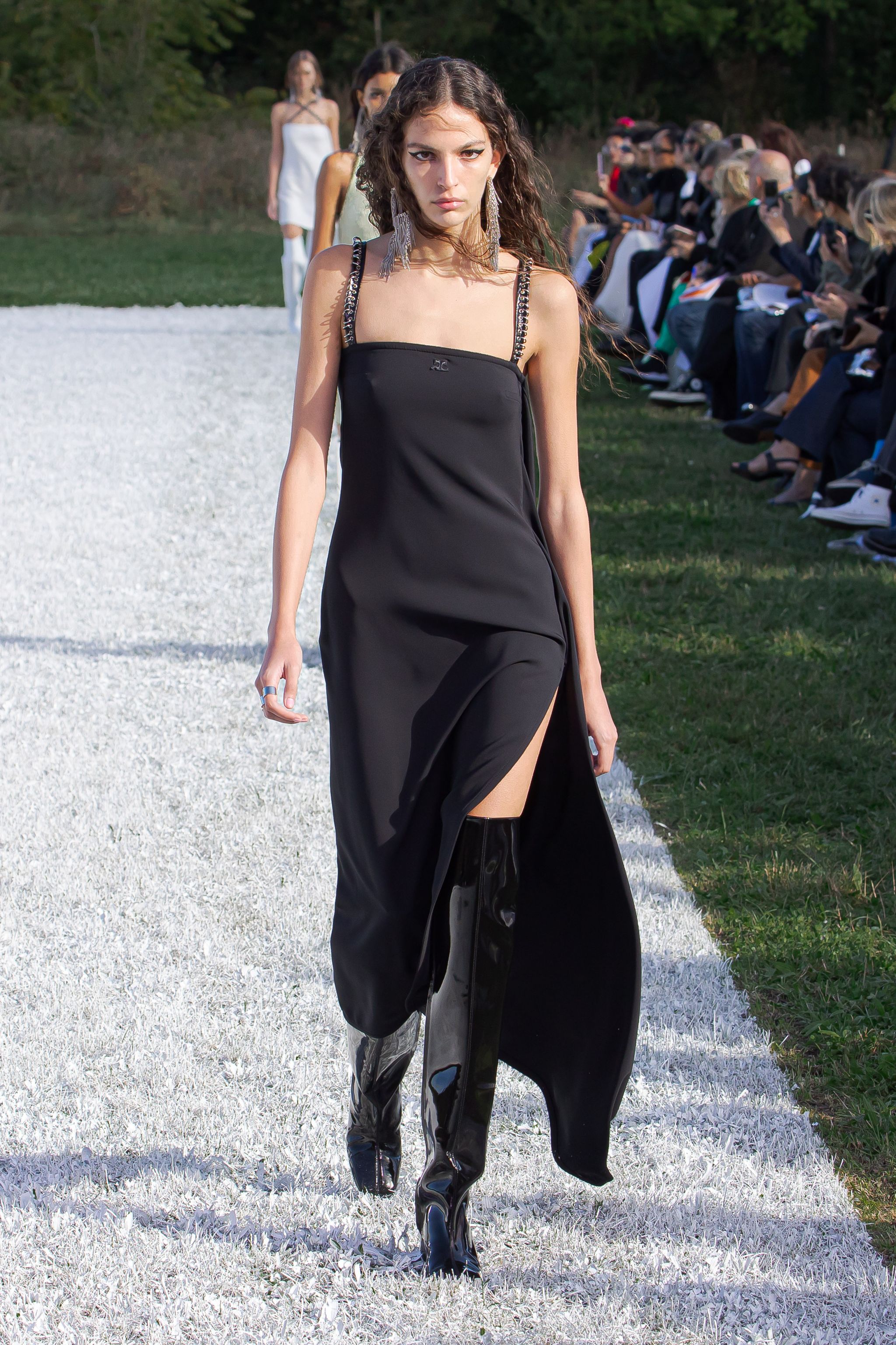 Elegant black Courrèges dress with unique straps and thigh-high patent boots on runway.