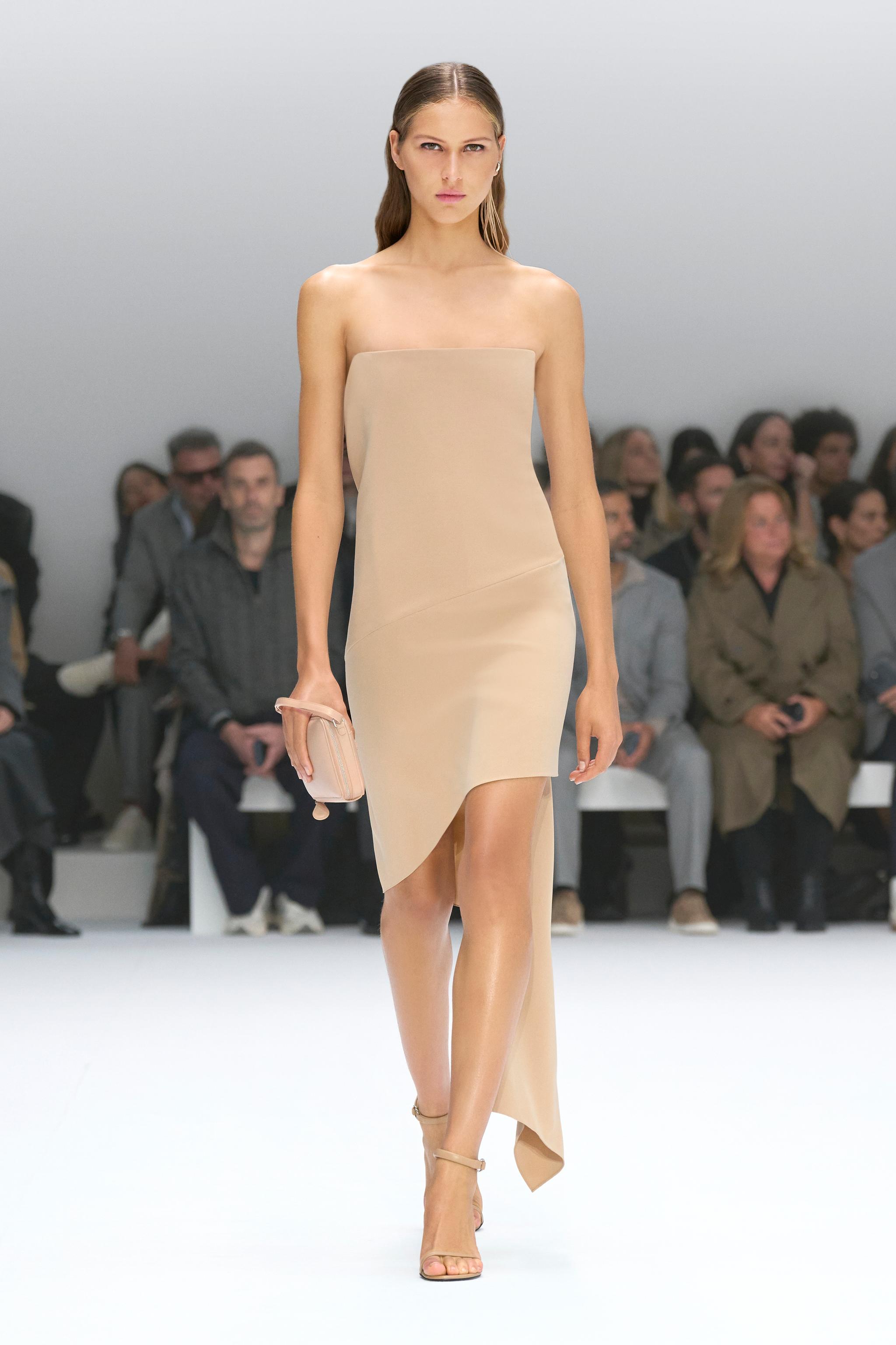 Elegant beige strapless dress by Courrèges showcased on a runway with audience in the background.