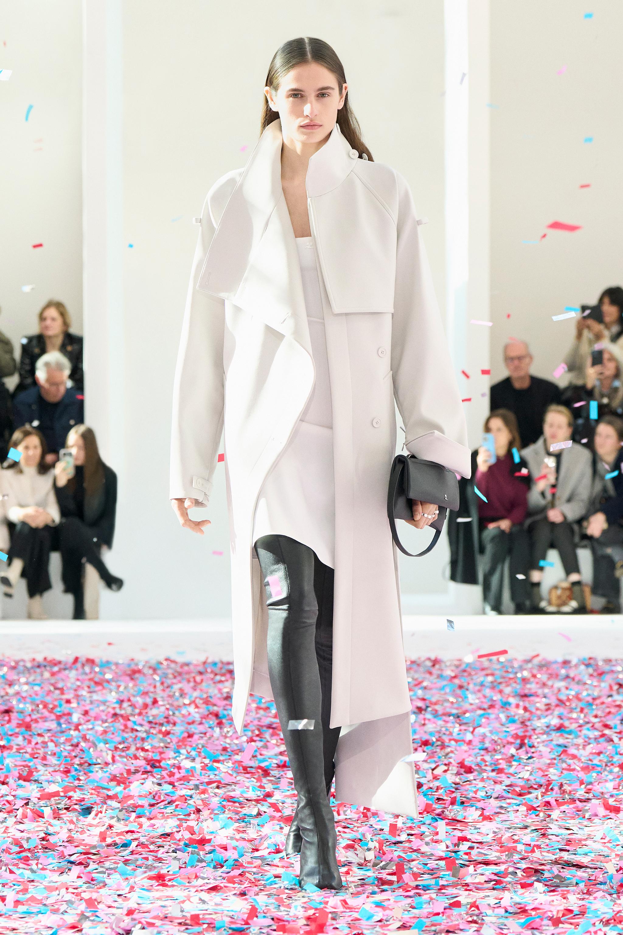 Elegant white trench coat by Courrèges showcased on runway with colorful confetti at fashion show.