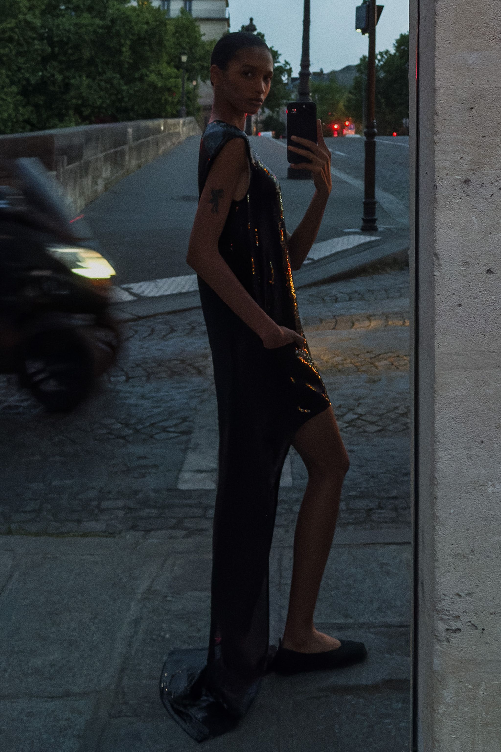 Elegant woman in a black sequin dress taking a mirror selfie on a city street for Courrèges.