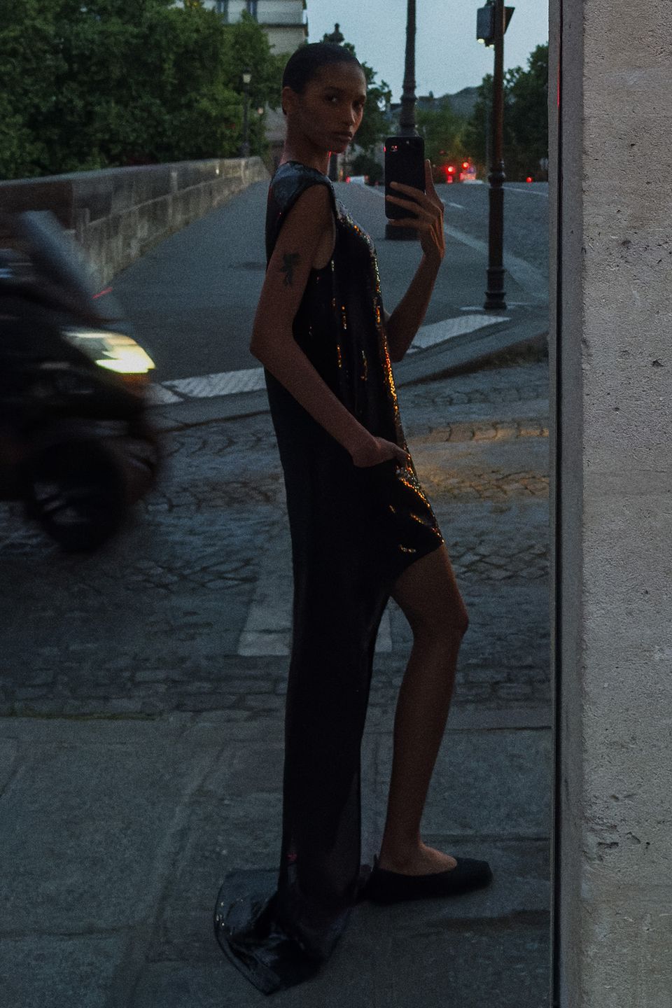 Elegant woman in a black sequin dress taking a mirror selfie on a city street for Courrèges.