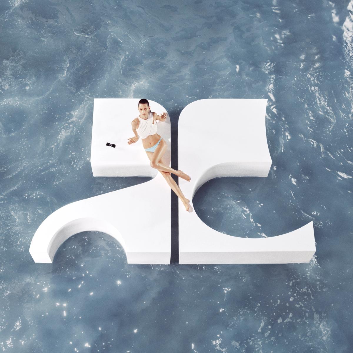 Elegant model in swimwear on Courreges logo sculpture in water, showcasing modern fashion.
