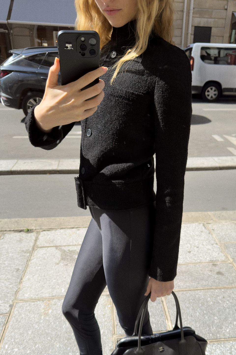 Elegant woman in black Courrèges fashion outfit taking a selfie outdoors on the sidewalk.