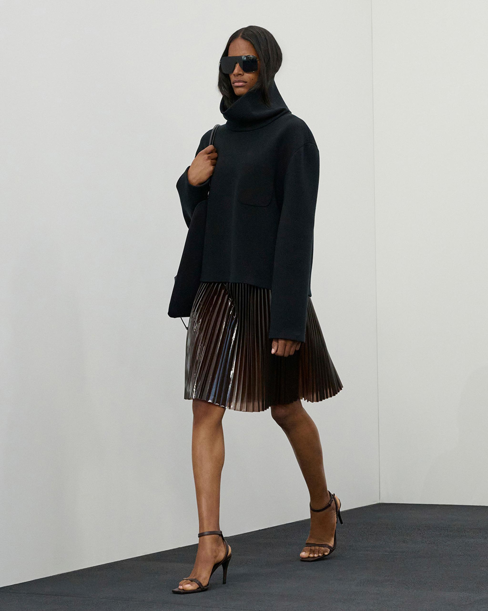Chic fashion model in black turtleneck, pleated skirt, and designer sunglasses on ecommerce site.
