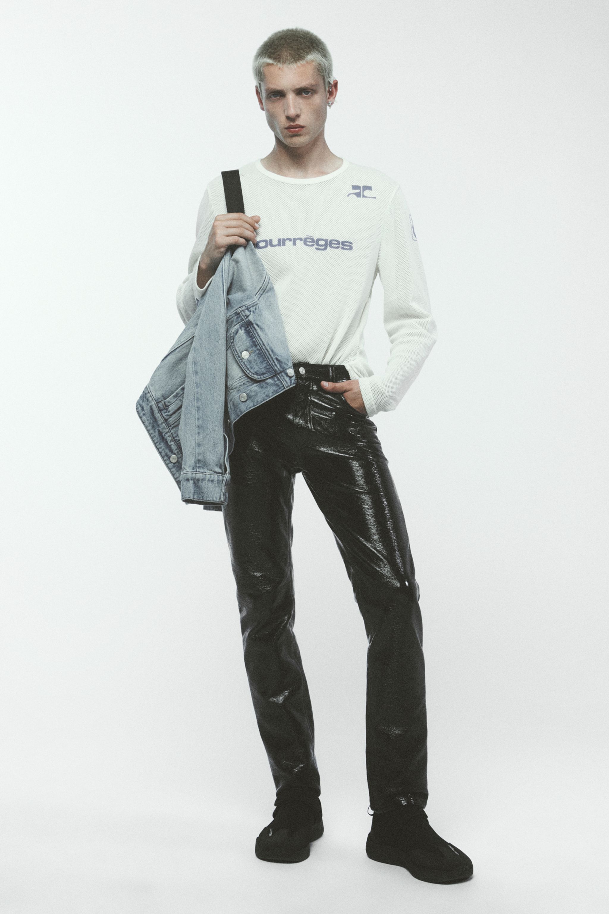 Model wearing Courrèges fashion, white sweater with logo, black patent pants, holding denim jacket, ecommerce style.