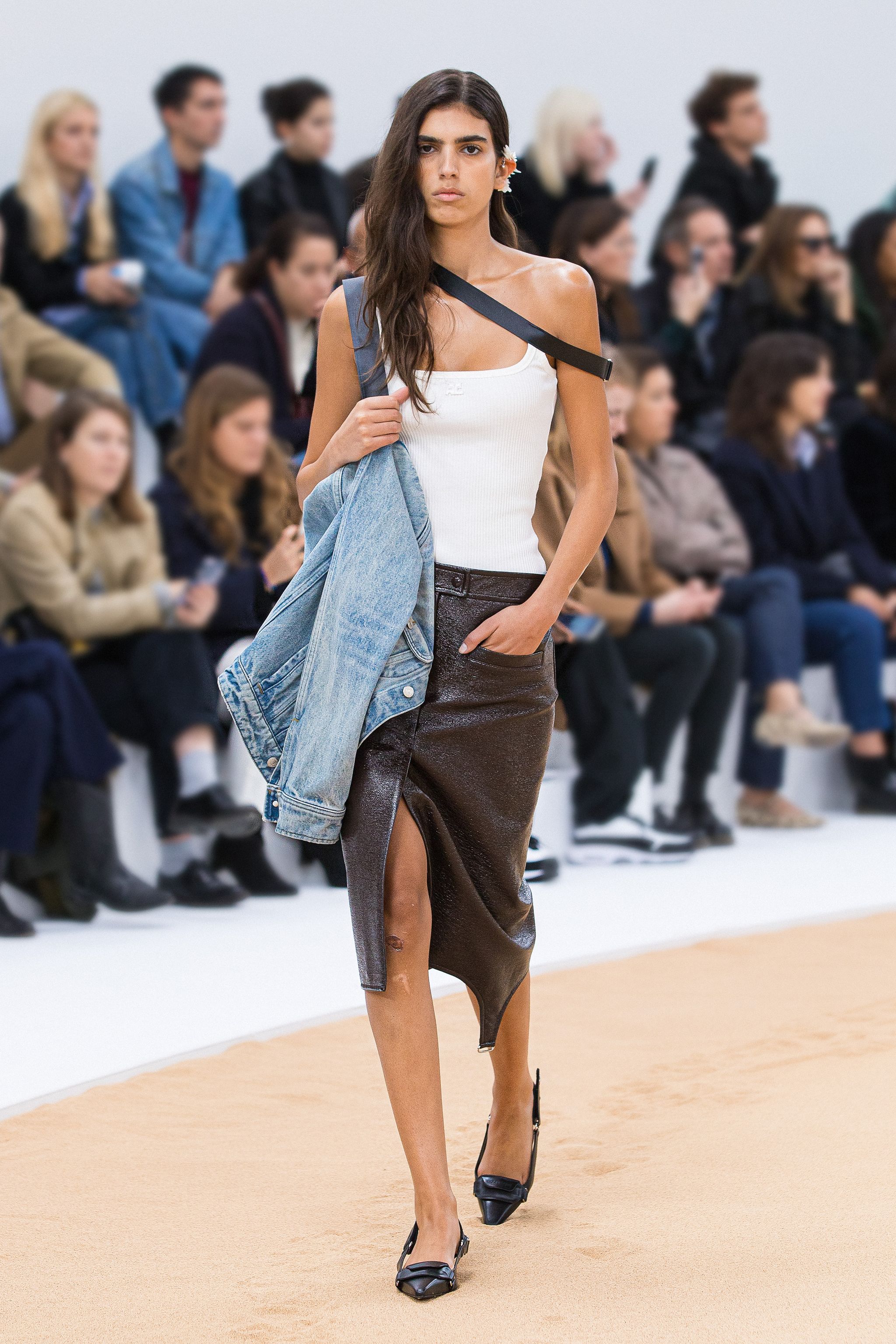 Elegant runway model wearing a white tank top, leather skirt, and carrying a denim jacket, Courrèges style.
