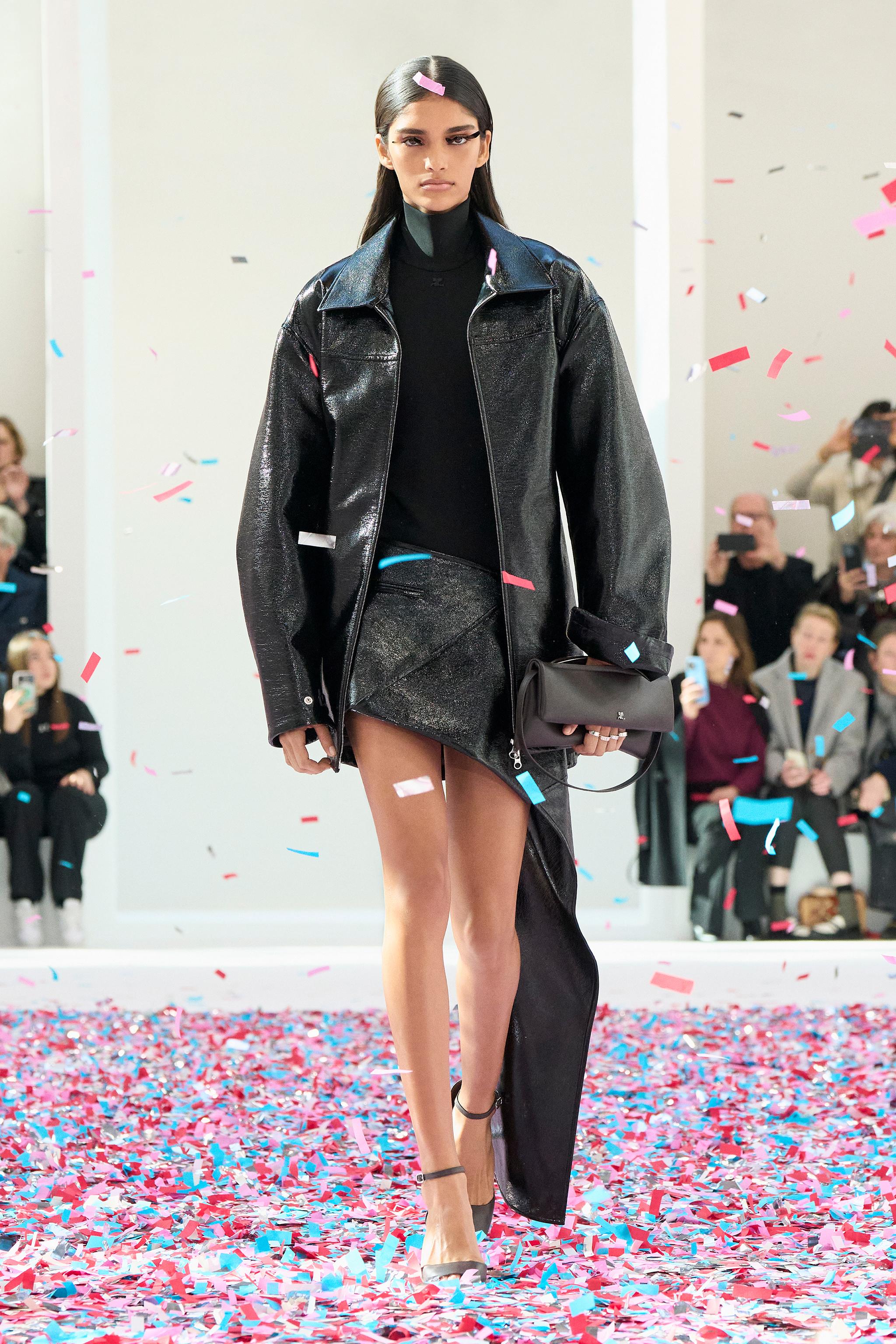 Elegant model in black leather jacket and skirt on runway, surrounded by colorful confetti, Courrèges fashion.