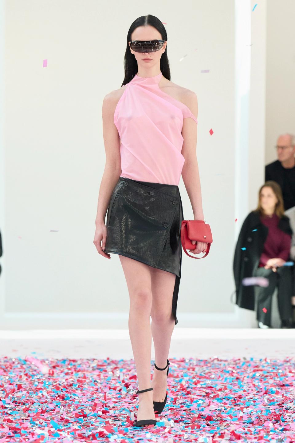 Elegant model wearing Courrèges fashion, pink top, black skirt, and accessories, on runway.