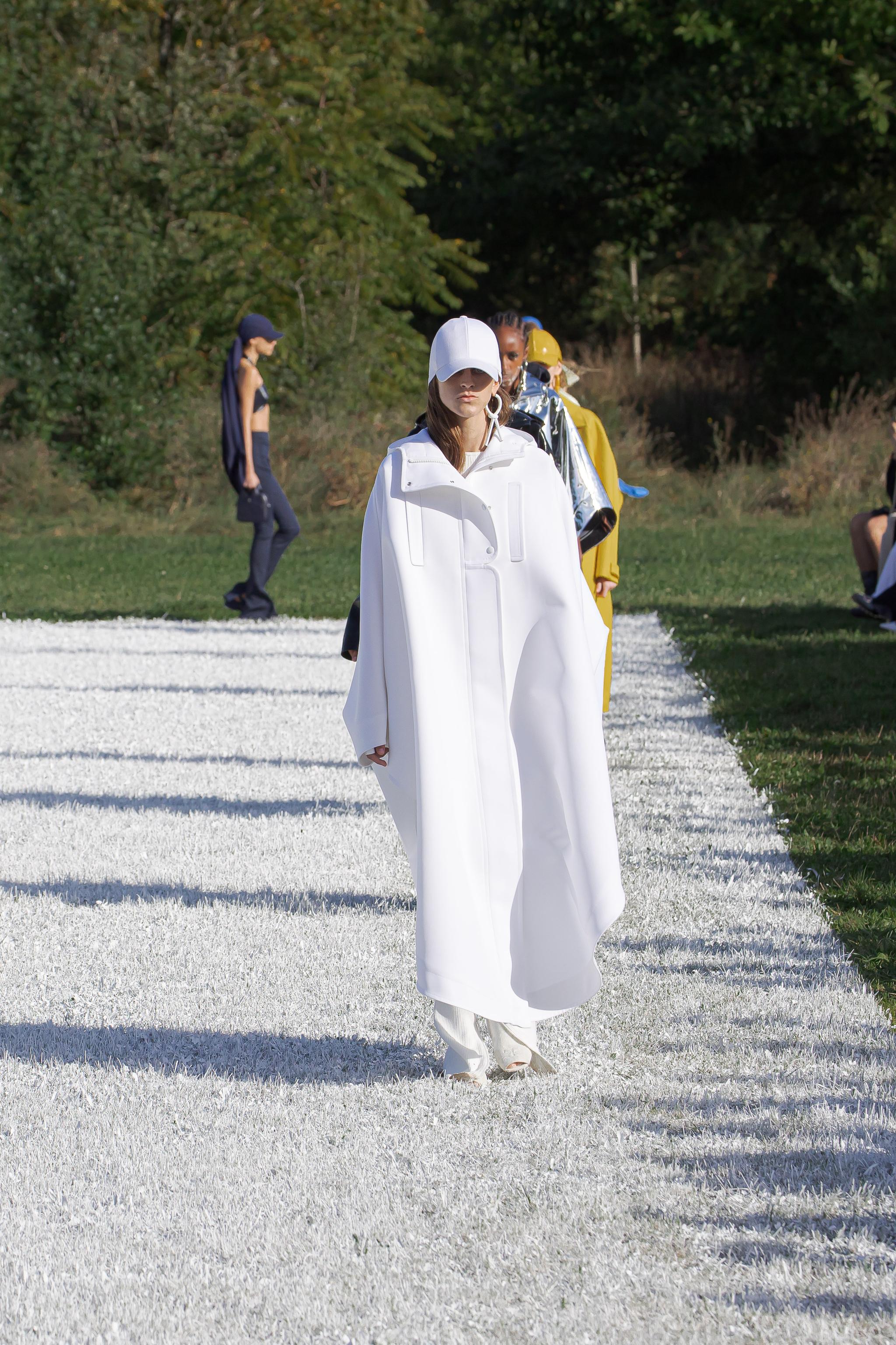 Elegant Courrèges model in a white futuristic coat and cap on a runway outdoors.