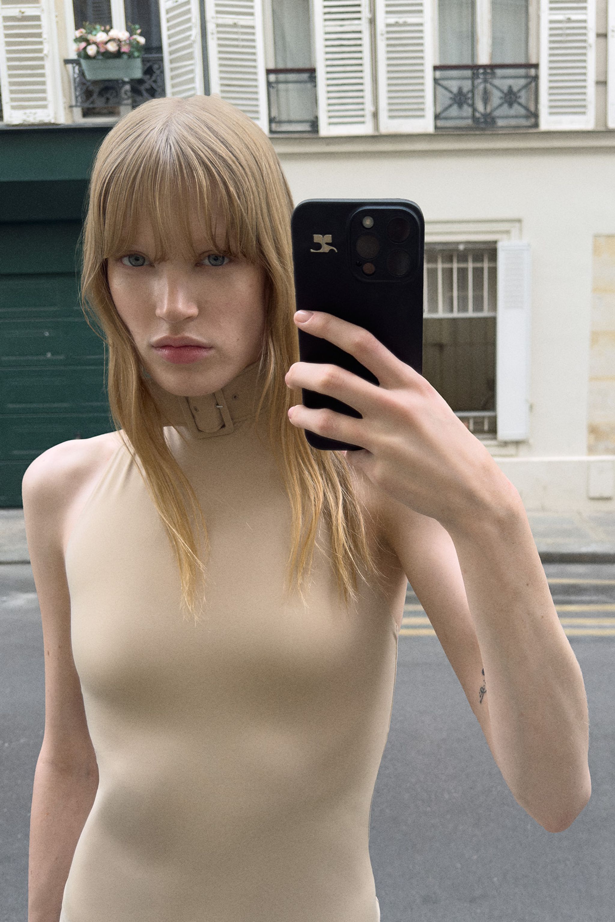 Fashion model wearing a beige top, taking a selfie on a city street for Courrèges.