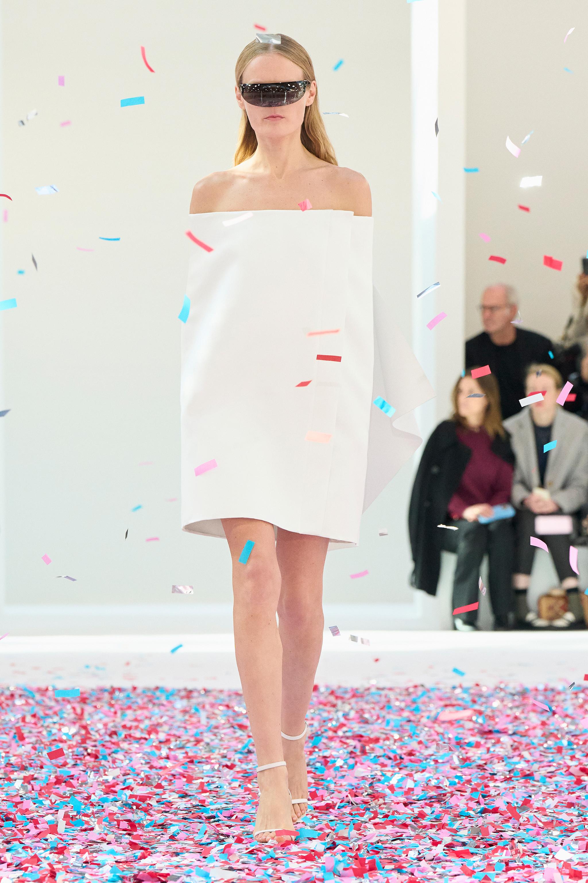 Elegant model in white Courrèges dress with futuristic sunglasses on a confetti-filled runway.