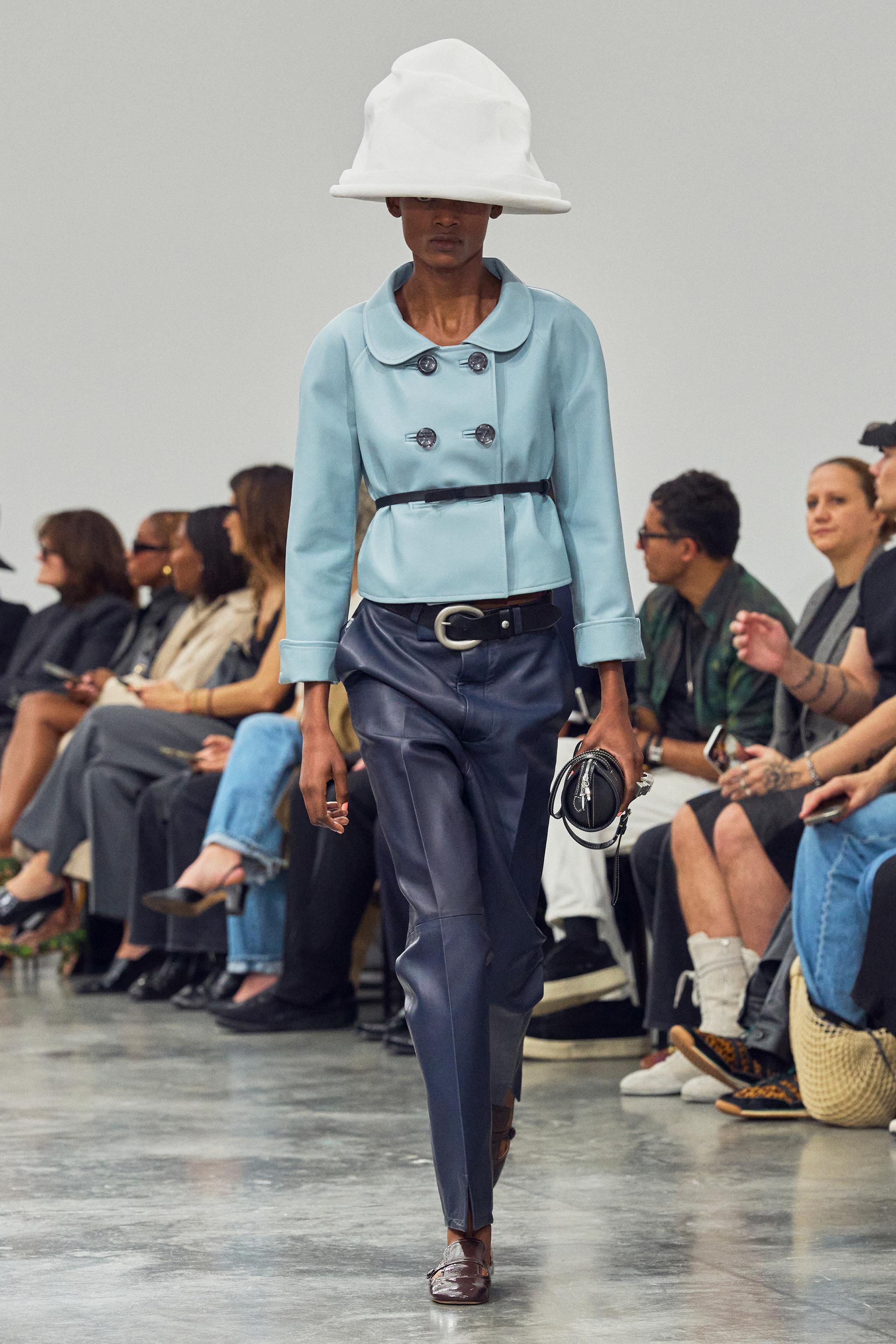 Elegant runway model wearing Courrèges fashion, light blue jacket, navy trousers, wide brim hat.