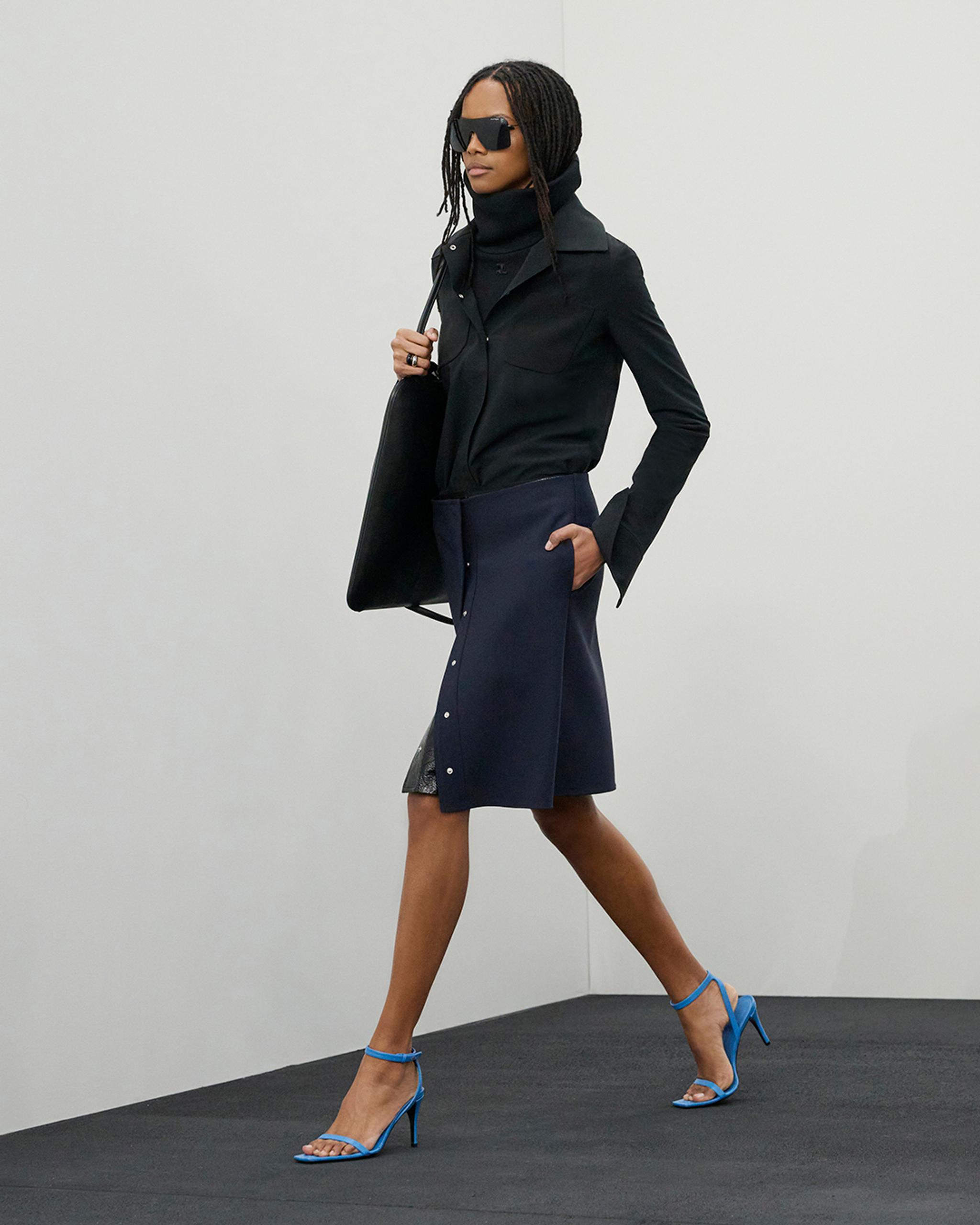 Elegant woman in black jacket and navy skirt with blue high heels, fashionably carrying a black bag for Courrèges.