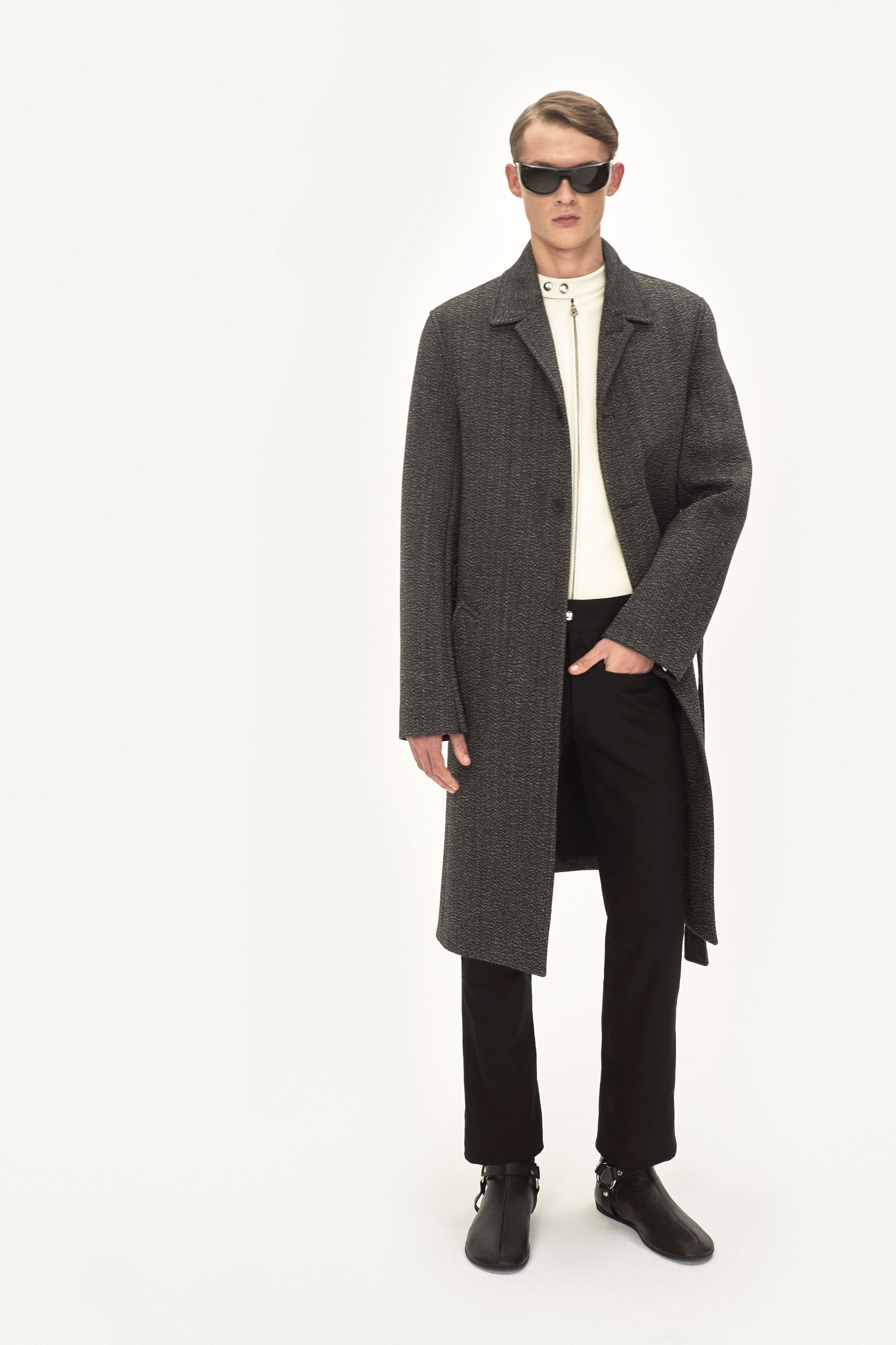 Elegant male fashion model wearing a long gray coat, white top, black trousers, and sunglasses on an ecommerce site for Courrèges.