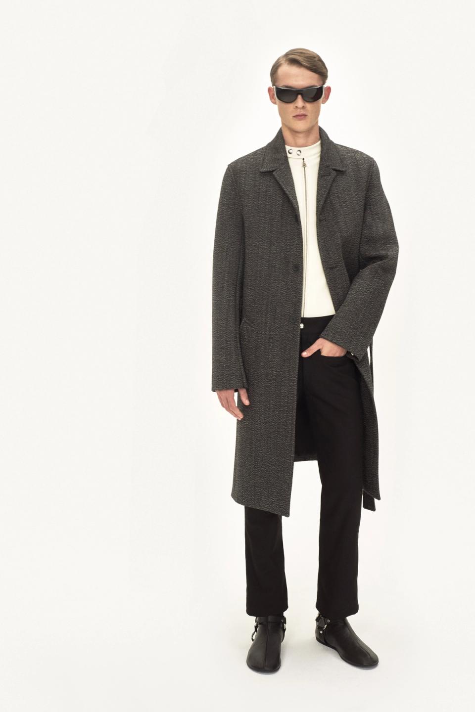 Elegant male fashion model wearing a long gray coat, white top, black trousers, and sunglasses on an ecommerce site for Courrèges.