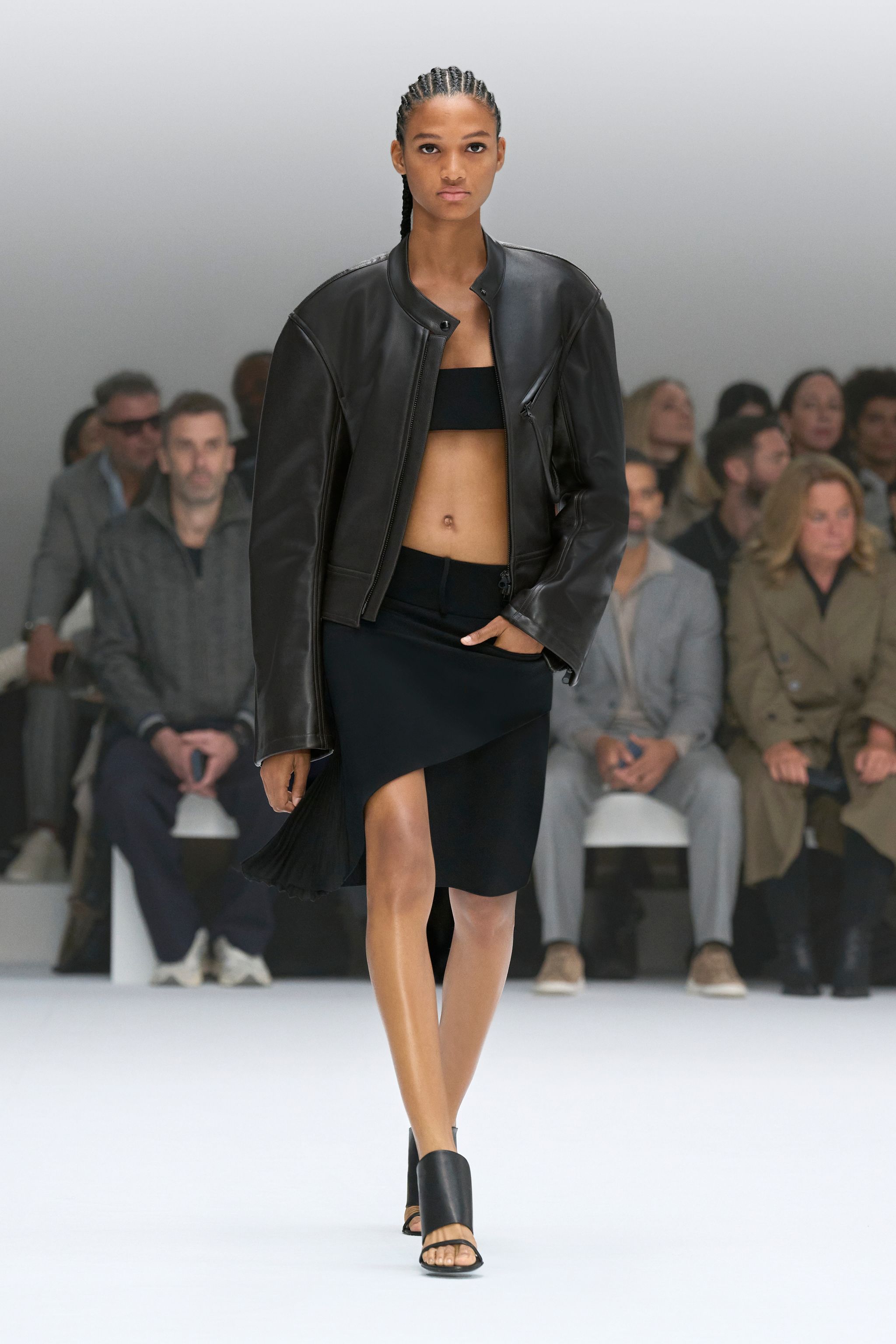 Elegant model in black leather jacket and skirt walk on runway for Courrèges fashion show.