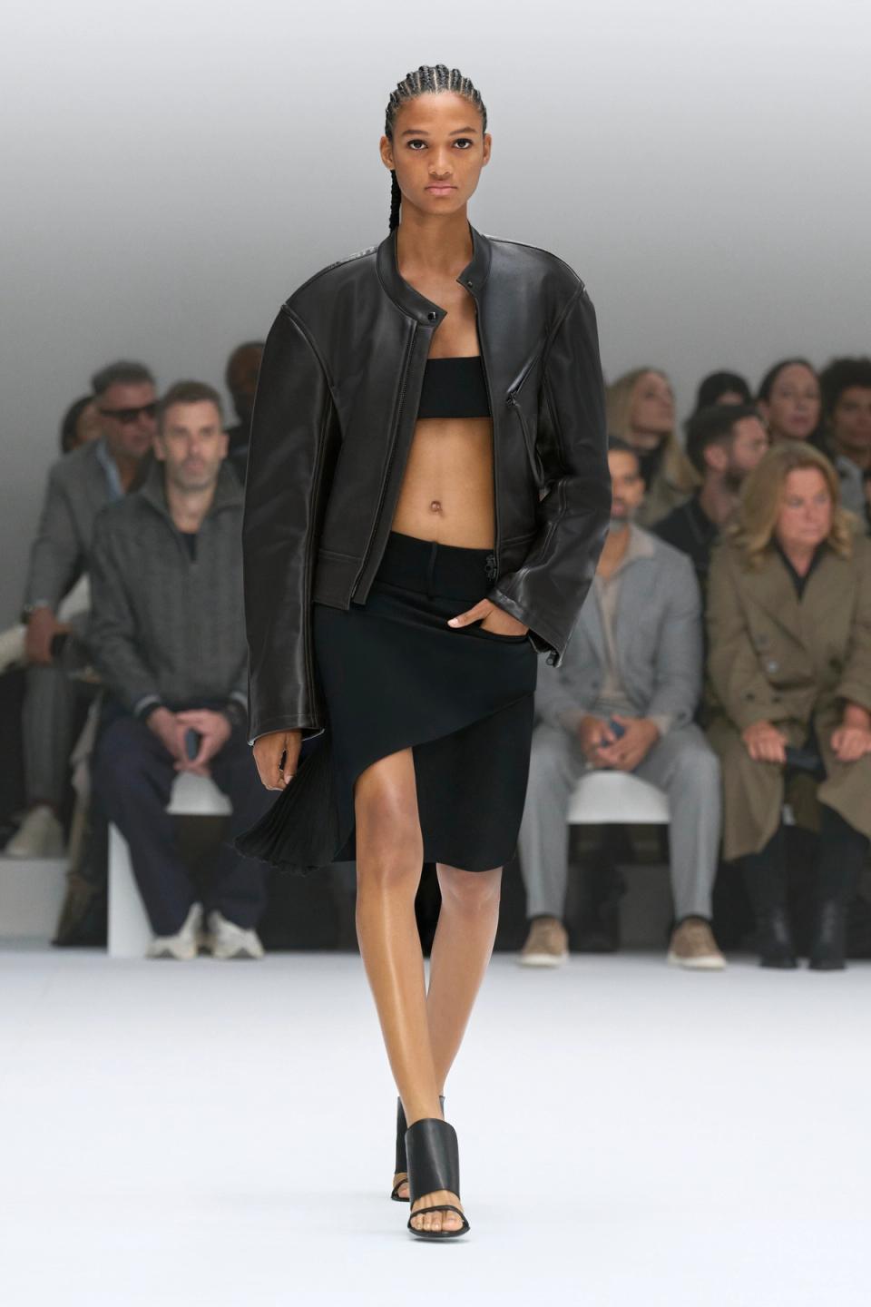 Elegant model in black leather jacket and skirt walk on runway for Courrèges fashion show.