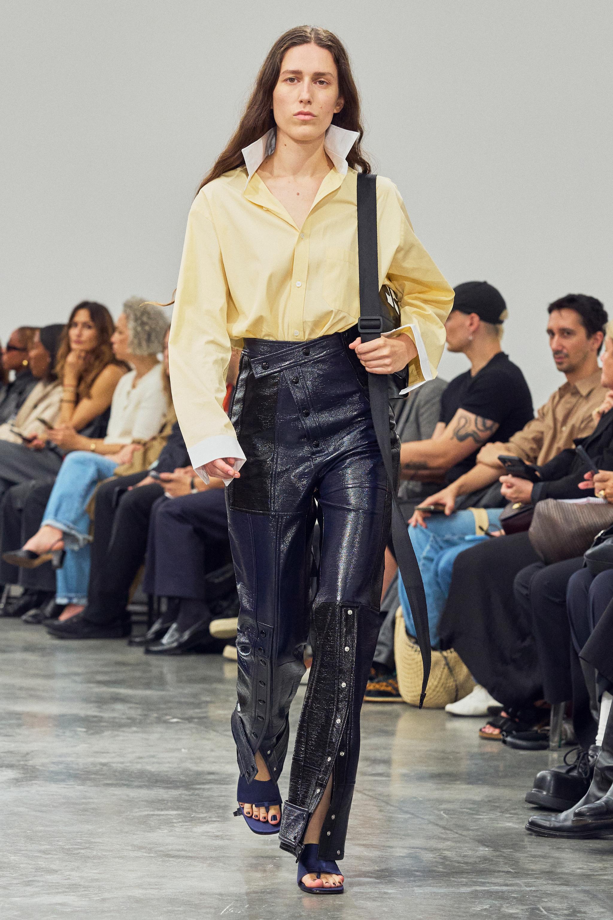 Elegant fashion runway look featuring a yellow shirt, high-waisted black leather pants, and a stylish black shoulder bag for Courrèges.