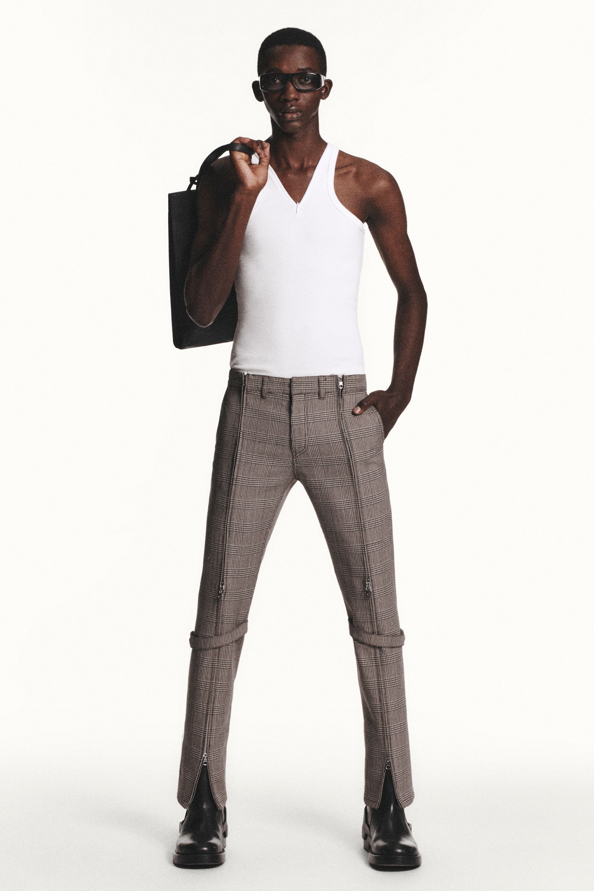 Elegant female model wearing Courrèges plaid pants, white top, black boots, and carrying a bag.
