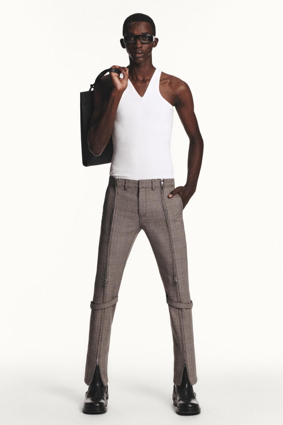 Elegant female model wearing Courrèges plaid pants, white top, black boots, and carrying a bag.