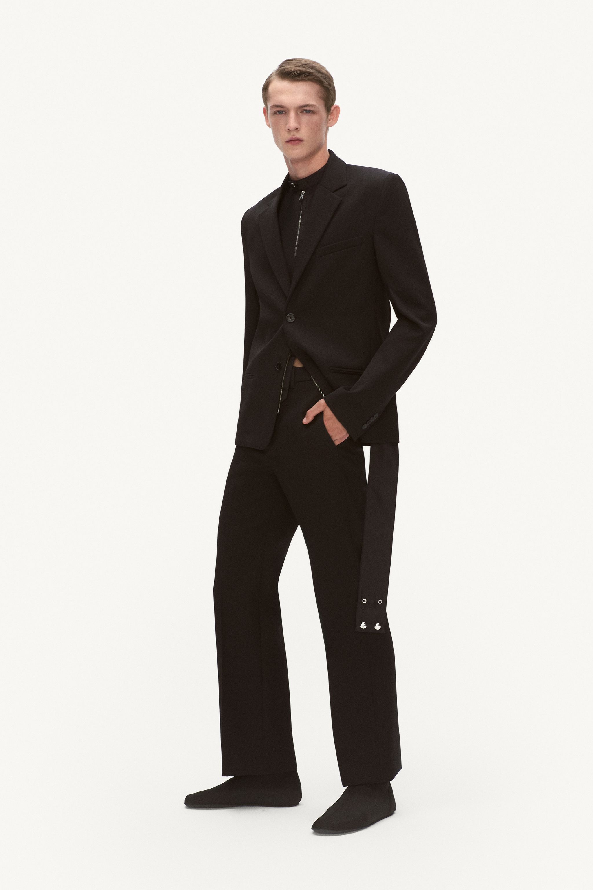 Elegant male model in black tailored Courrèges suit and footwear on a white backdrop.