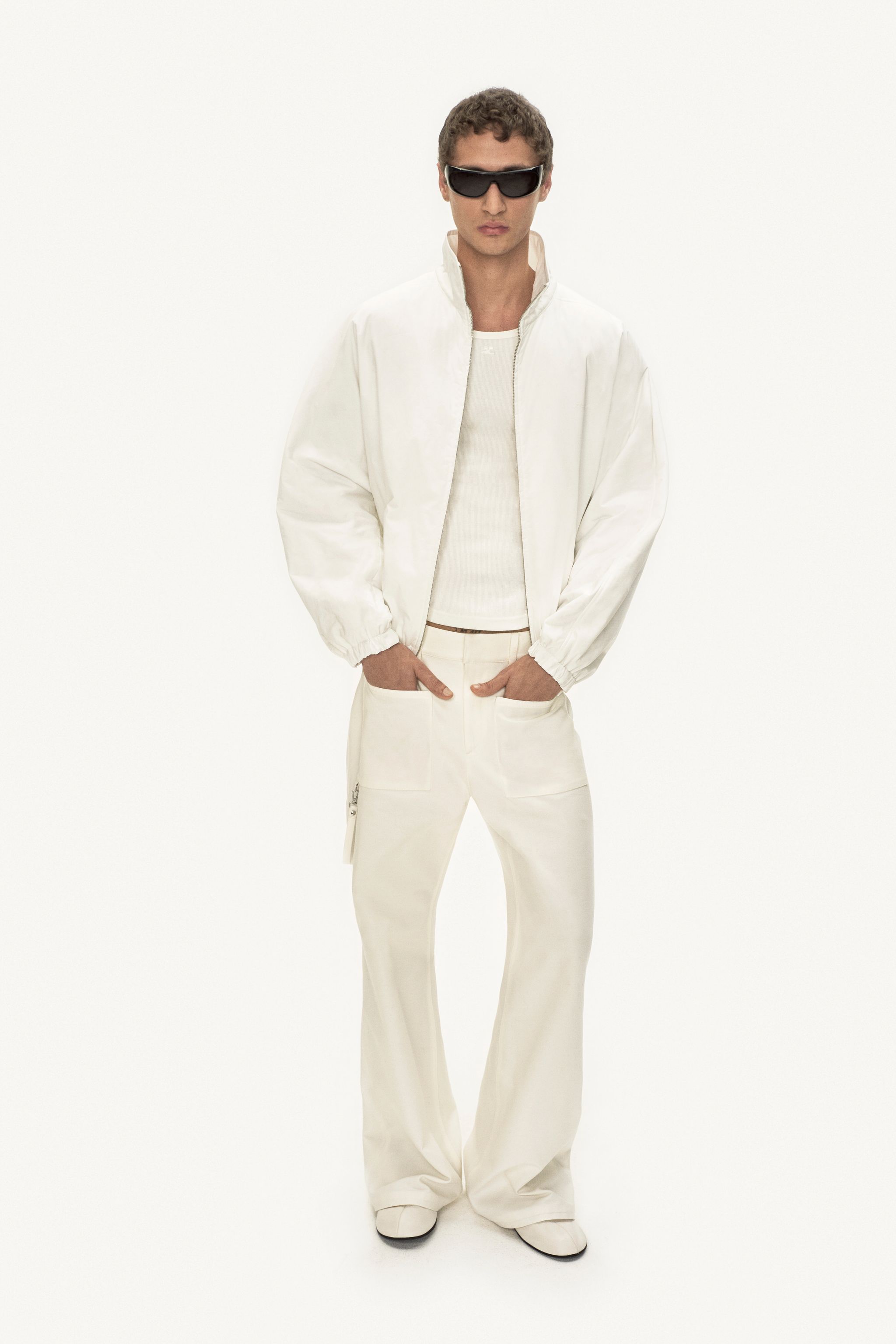 Elegant male model in white Courrèges fashion, wearing stylish sunglasses on white background.