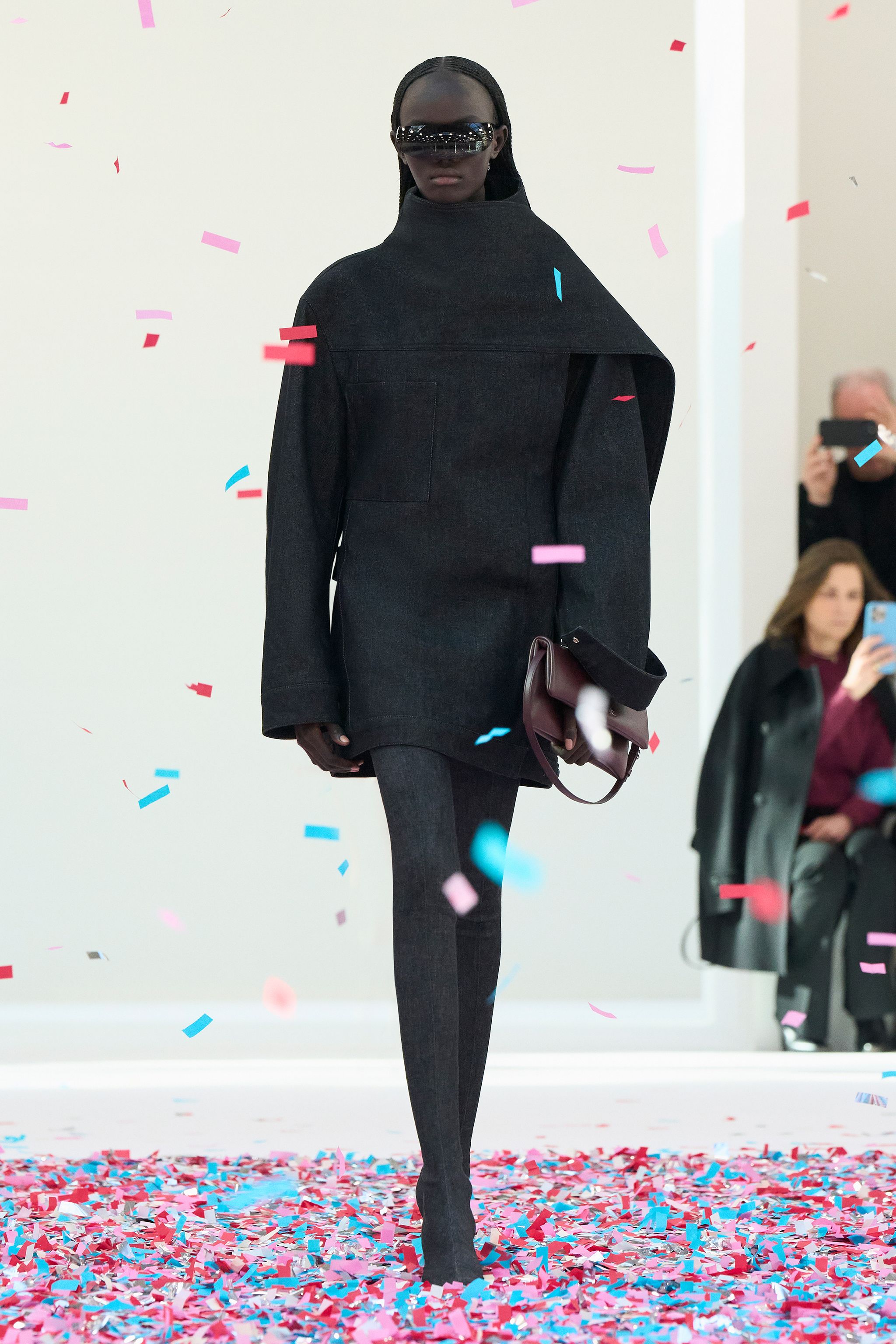 Elegant black oversized coat with futuristic sunglasses, flowing through confetti at a fashion show.