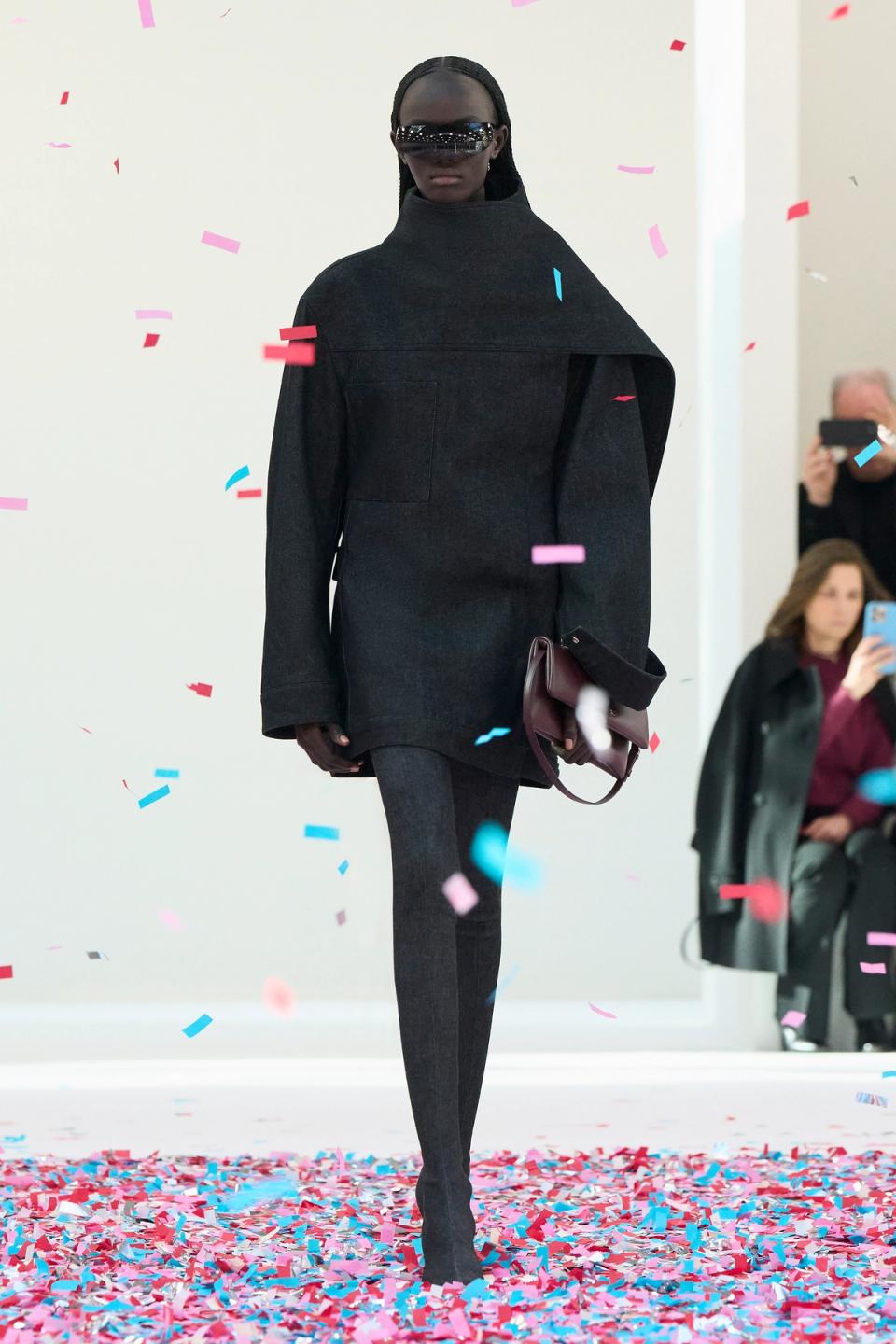 Elegant black oversized coat with futuristic sunglasses, flowing through confetti at a fashion show.