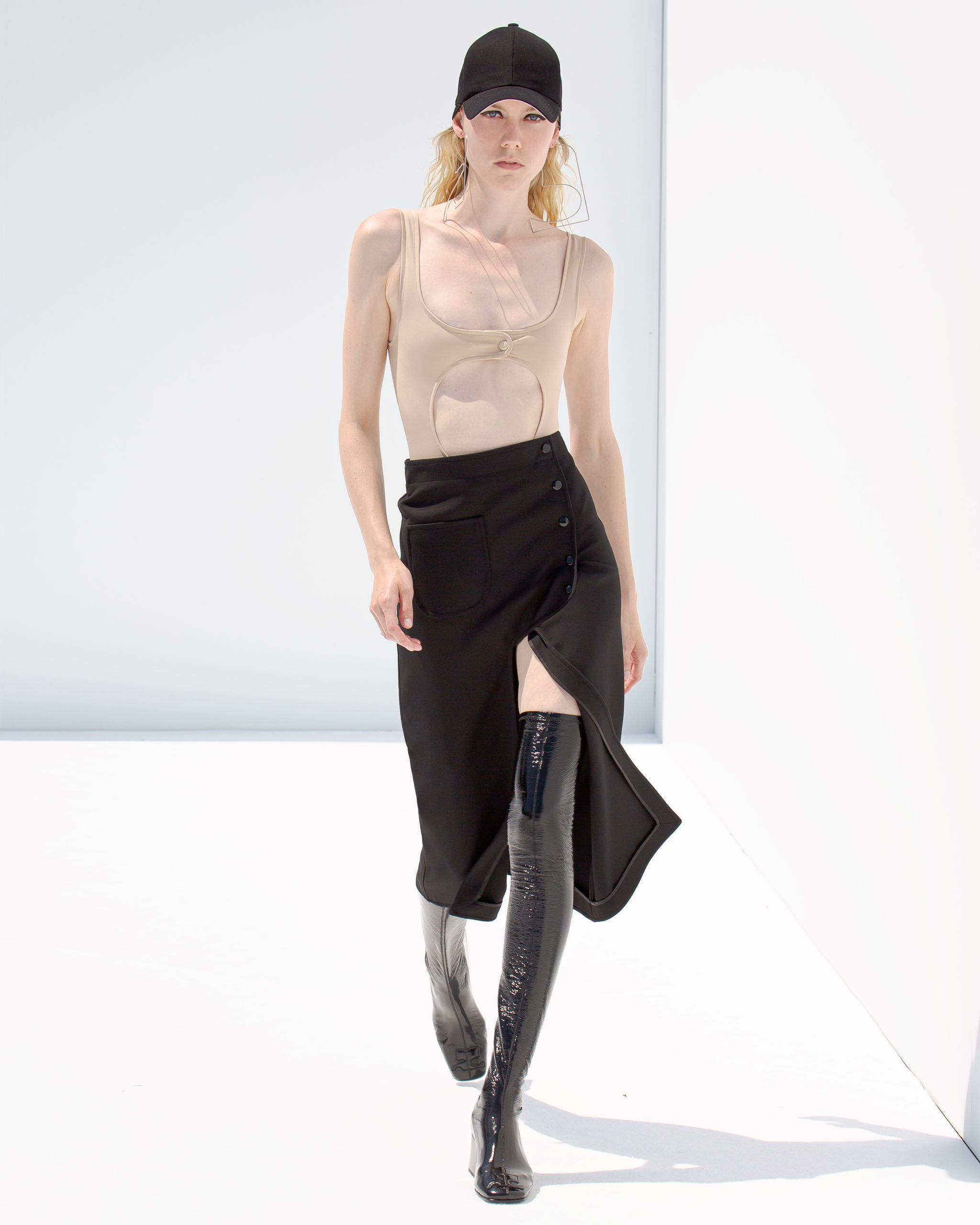 Elegant Courrèges fashion model wearing a beige cut-out top, black skirt, and shiny thigh-high boots.