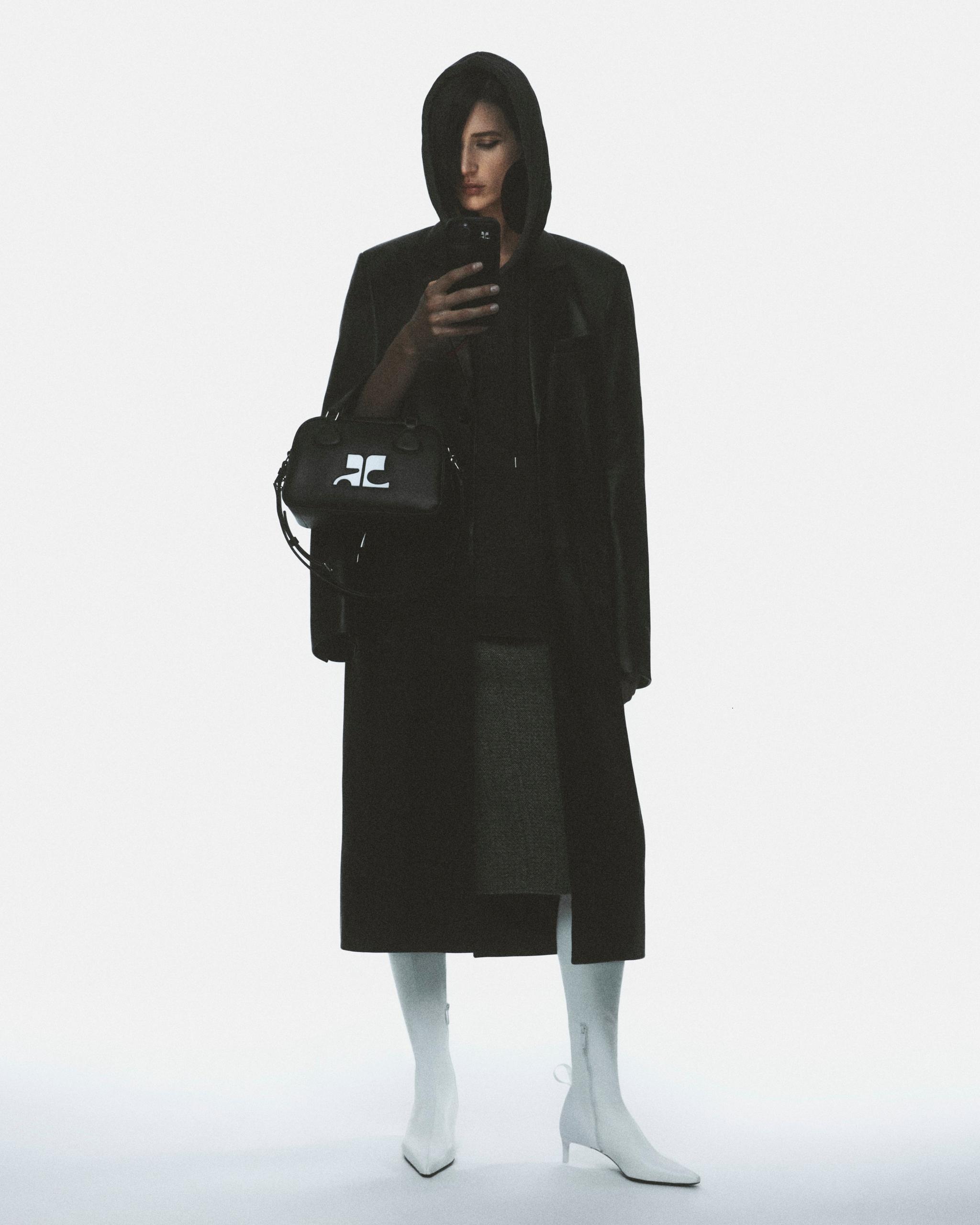 Elegant woman in black coat with hood, holding a Courrèges handbag in a fashion shoot.
