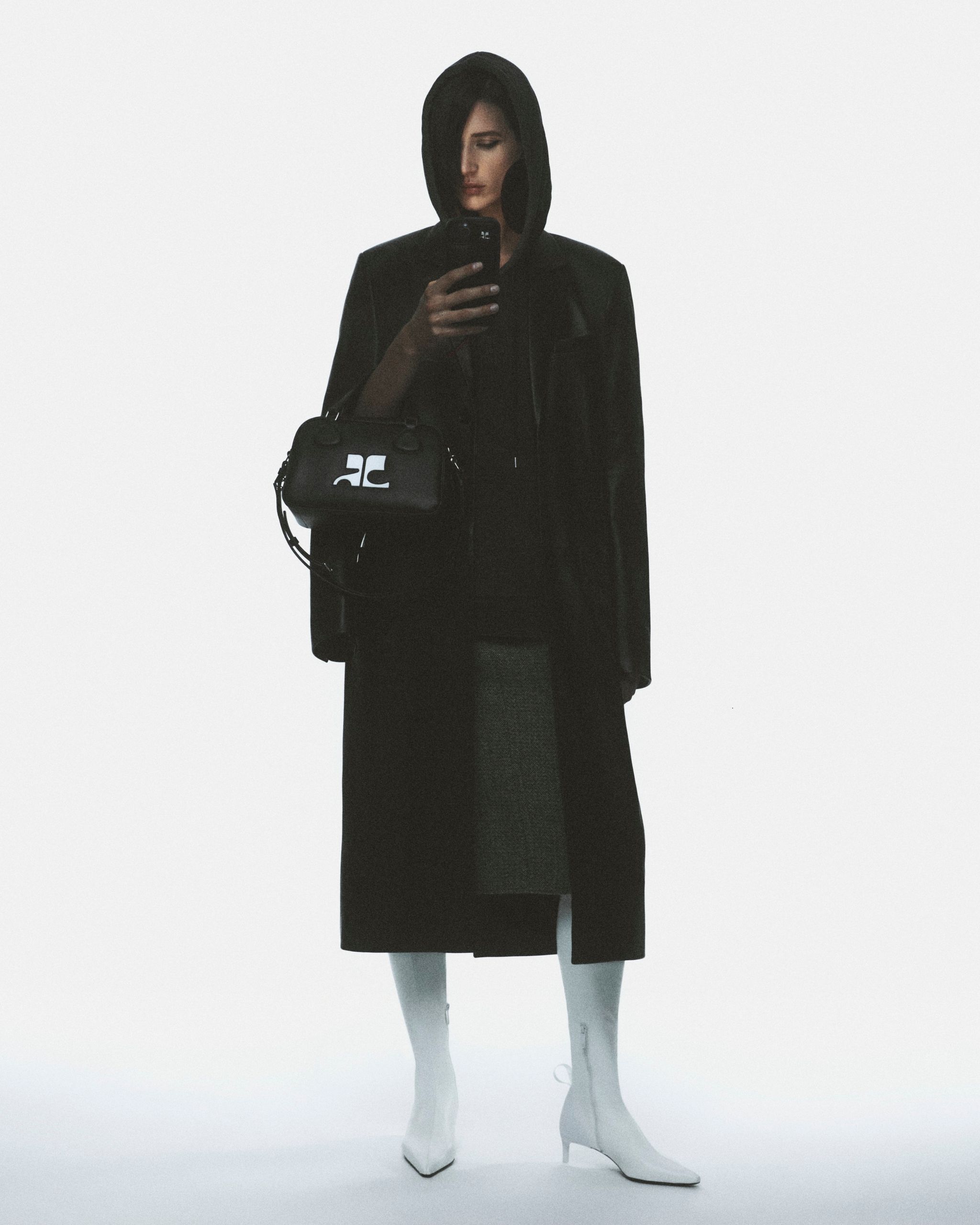 Femme élégante en noir avec capuche, portant un sac Courrèges, sur un site e-commerce.