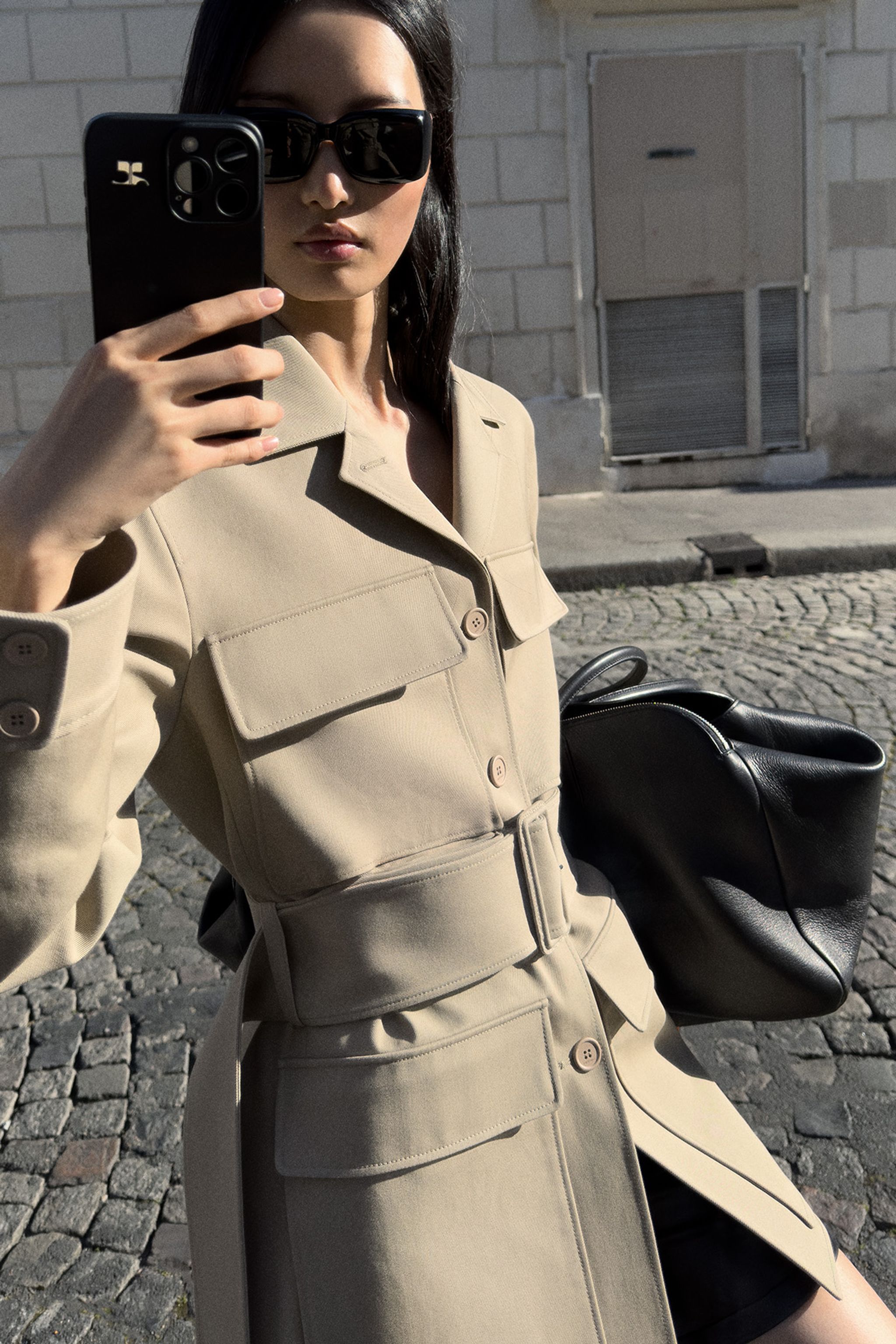 Chic woman in beige trench coat and black sunglasses taking a selfie on a city street for Courrèges.