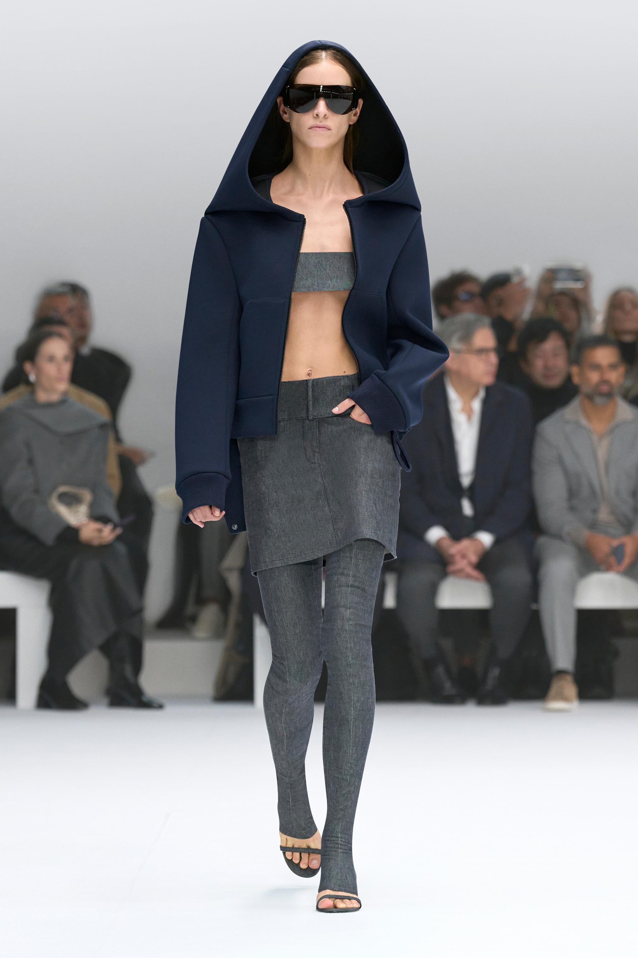 Elegant Courrèges fashion runway model in a navy hooded jacket, crop top, gray skirt, and leggings.