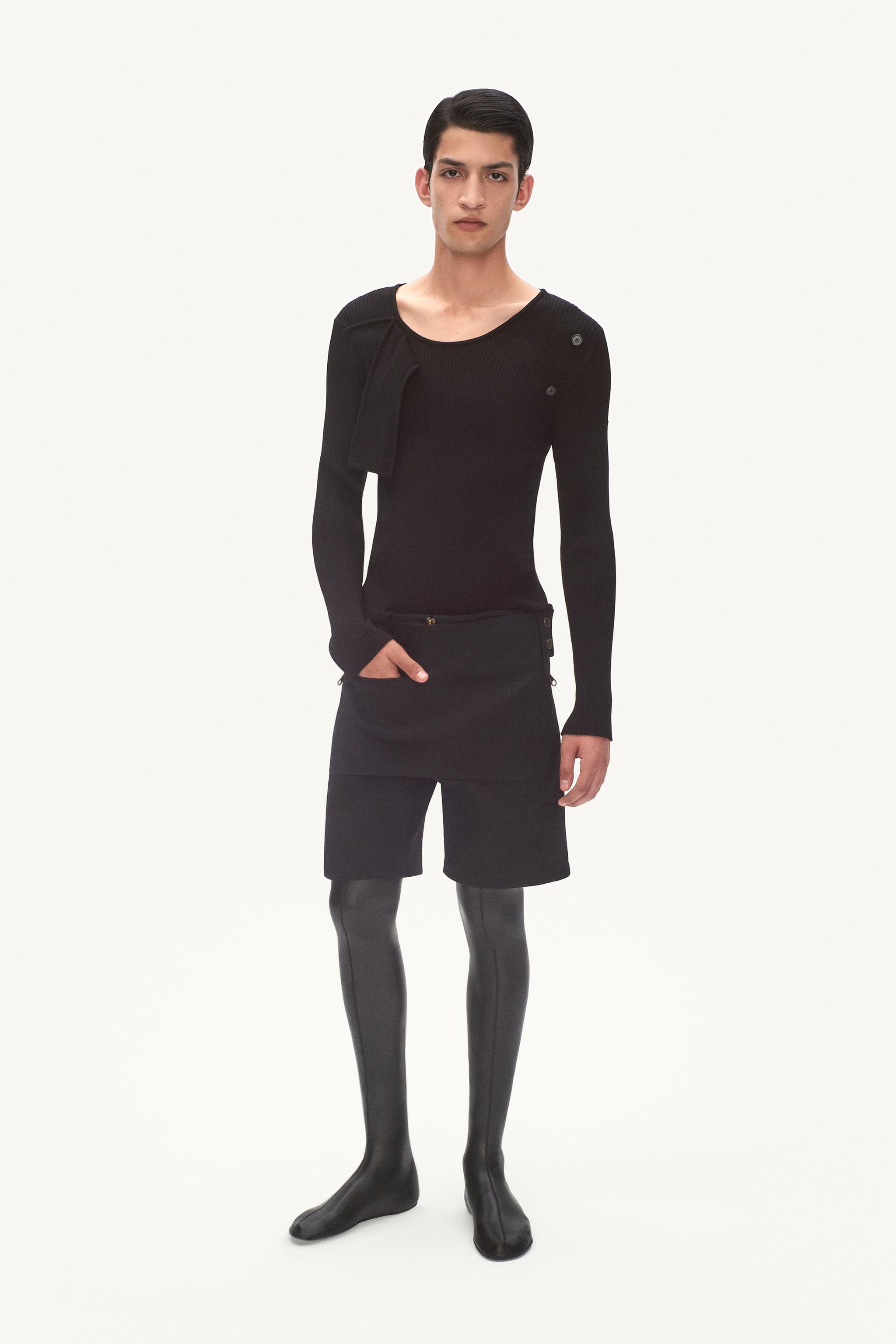 Elegant male model in black sweater and shorts, showcasing Courrèges fashion on minimalist background.