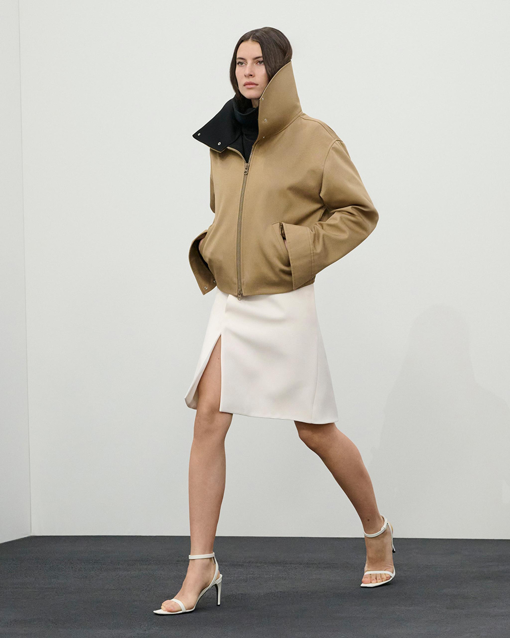 Elegant woman in Courrèges outfit with beige jacket, white skirt, and heels, on ecommerce site.