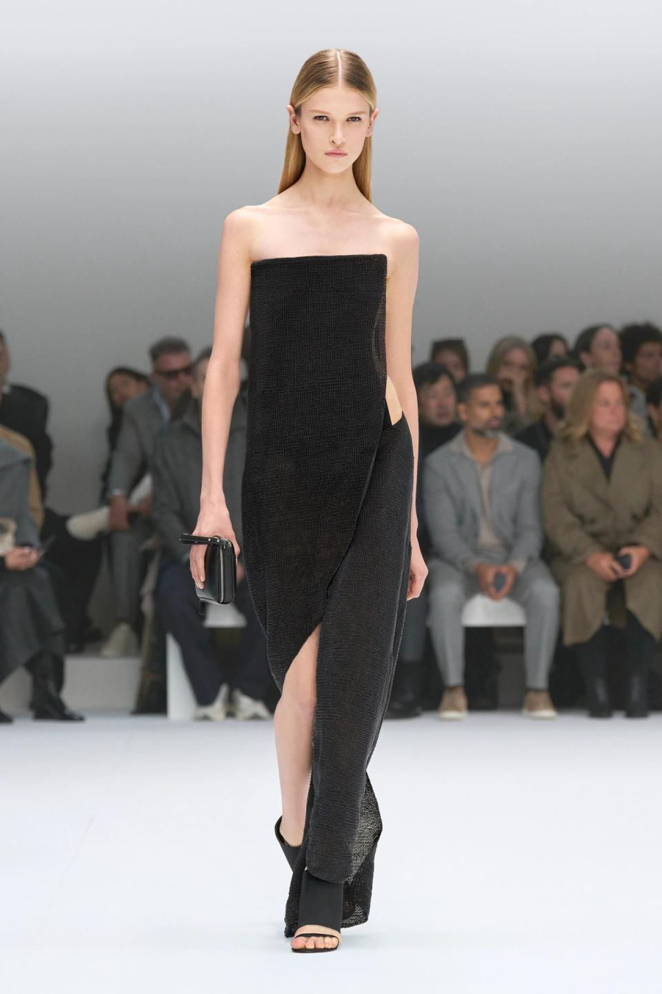 Elegant black strapless dress by Courrèges showcased on runway with audience in background.