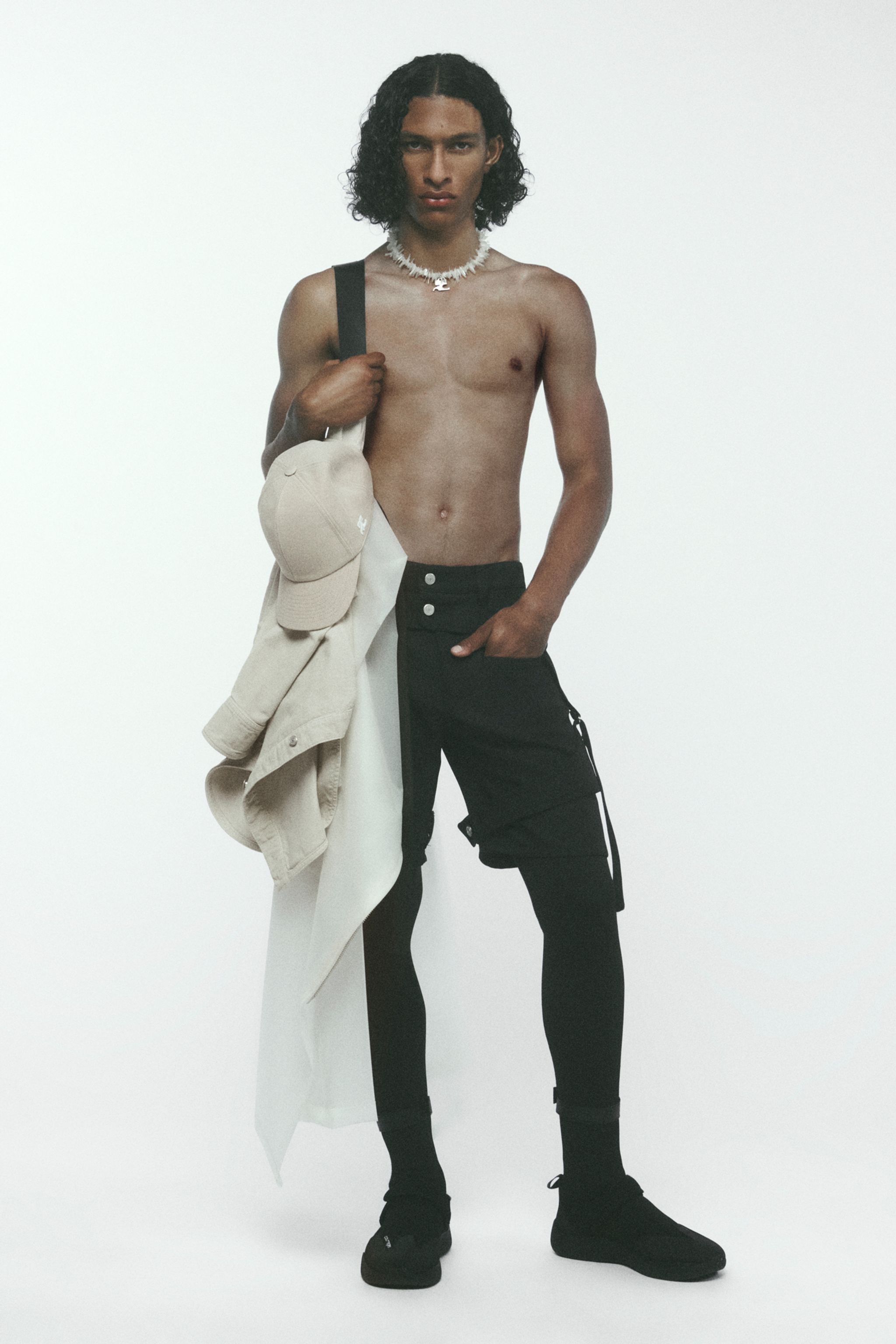 Male model wearing black cargo pants, holding beige jacket and cap, representing Courrèges fashion.