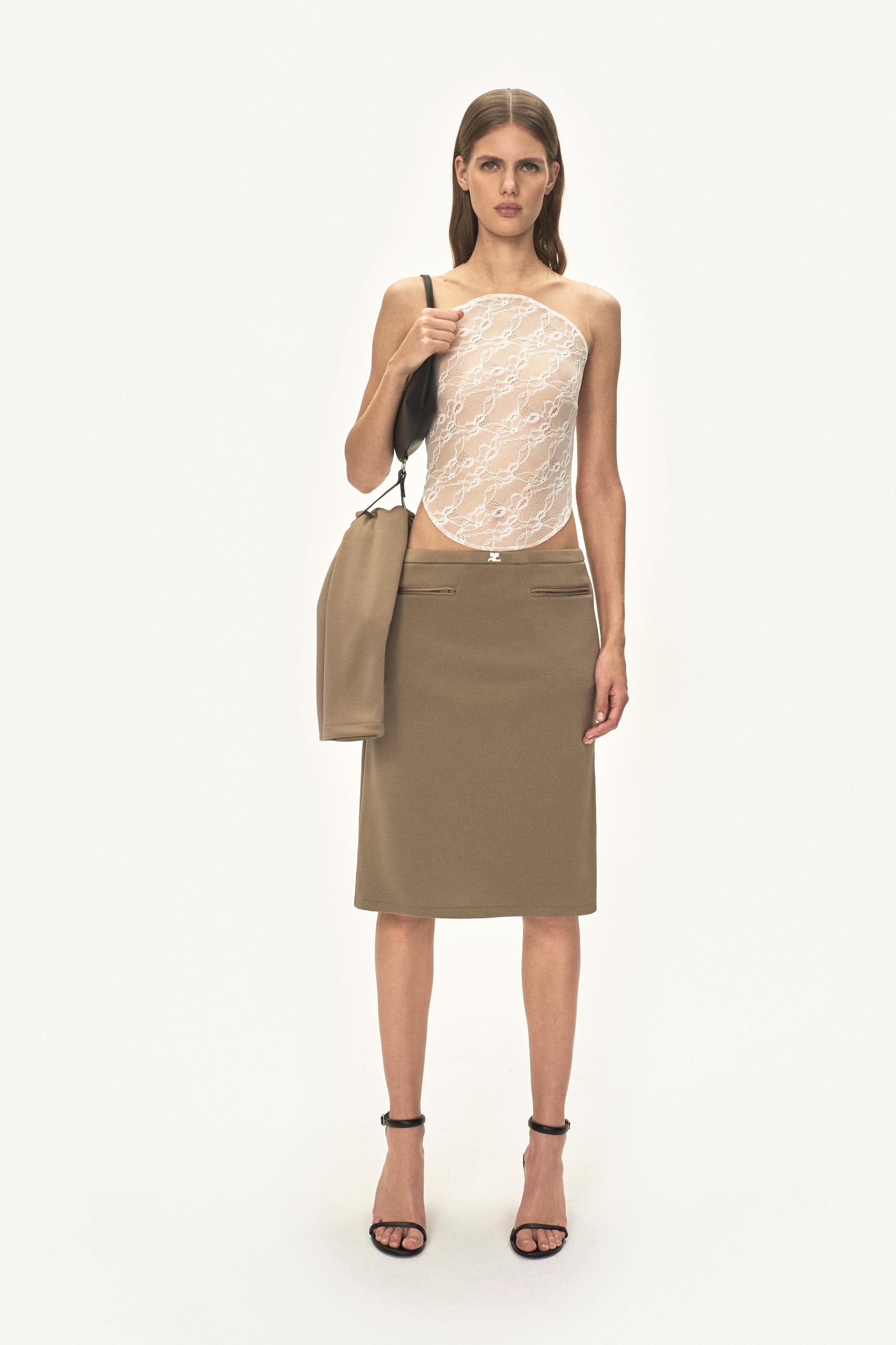Modèle féminin élégant portant un haut en dentelle Courrèges et une jupe beige, accessoirisé d'un sac noir.