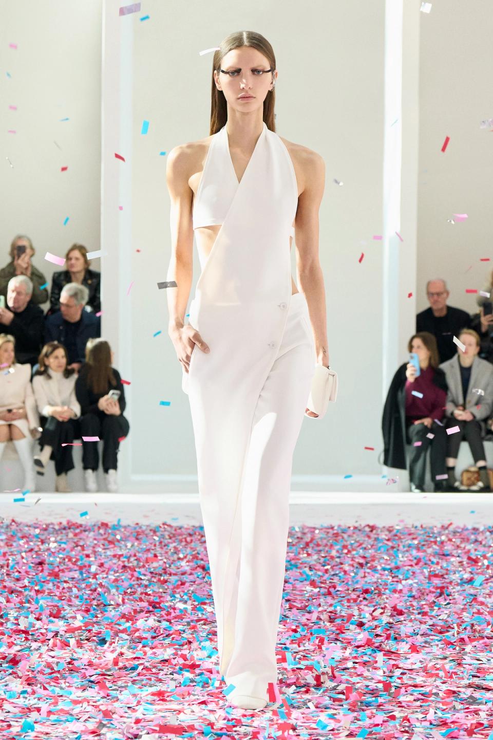 Elegant white modern jumpsuit by Courrèges showcased on a runway with colorful confetti.