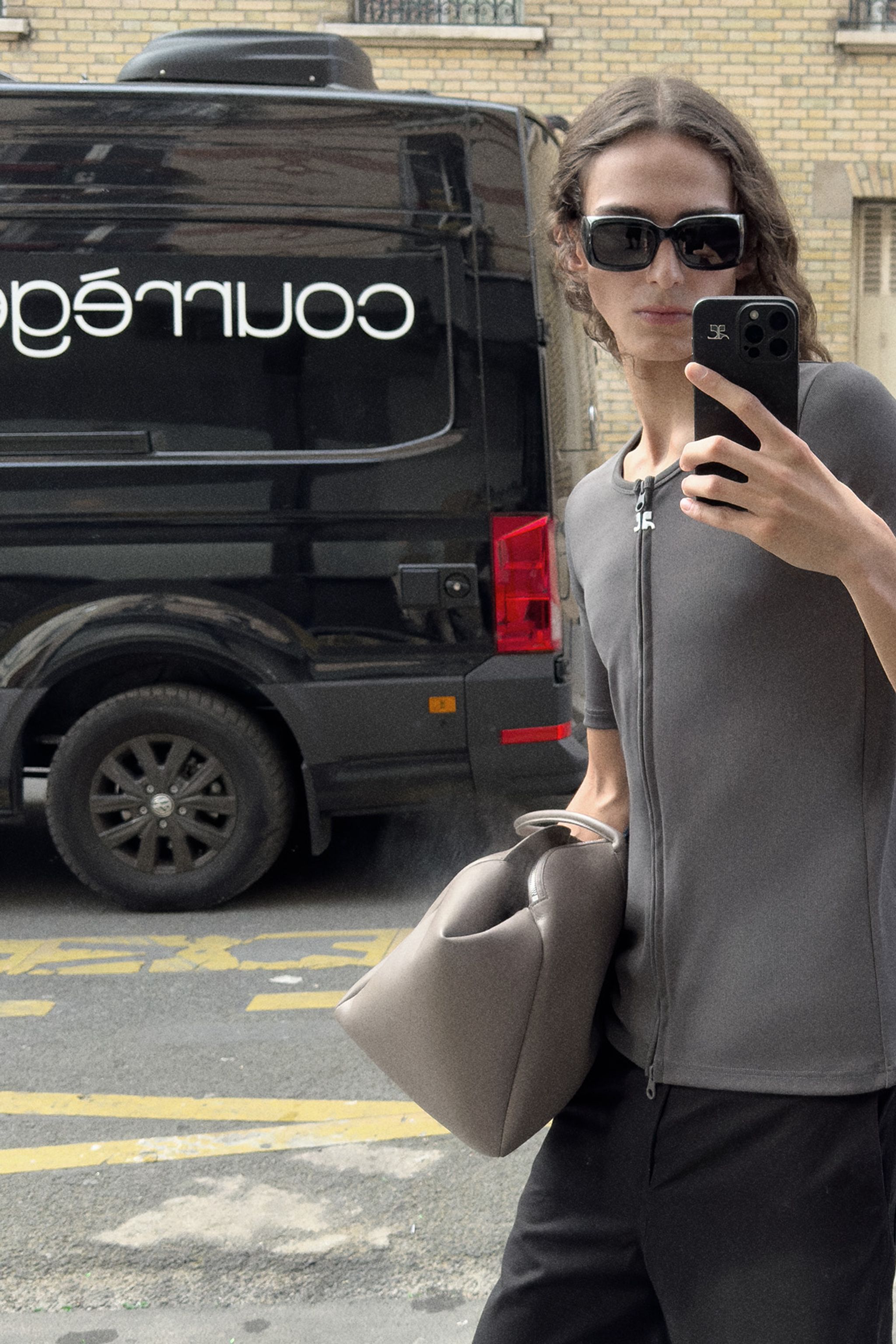 Elegant woman in gray jacket with handbag, taking mirror selfie on urban street, Courrèges style.