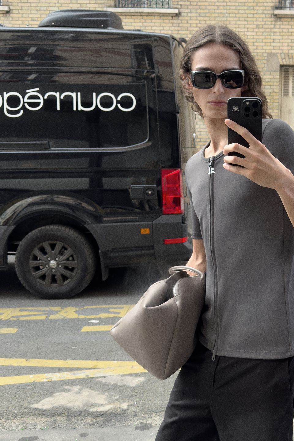 Elegant woman in gray jacket with handbag, taking mirror selfie on urban street, Courrèges style.