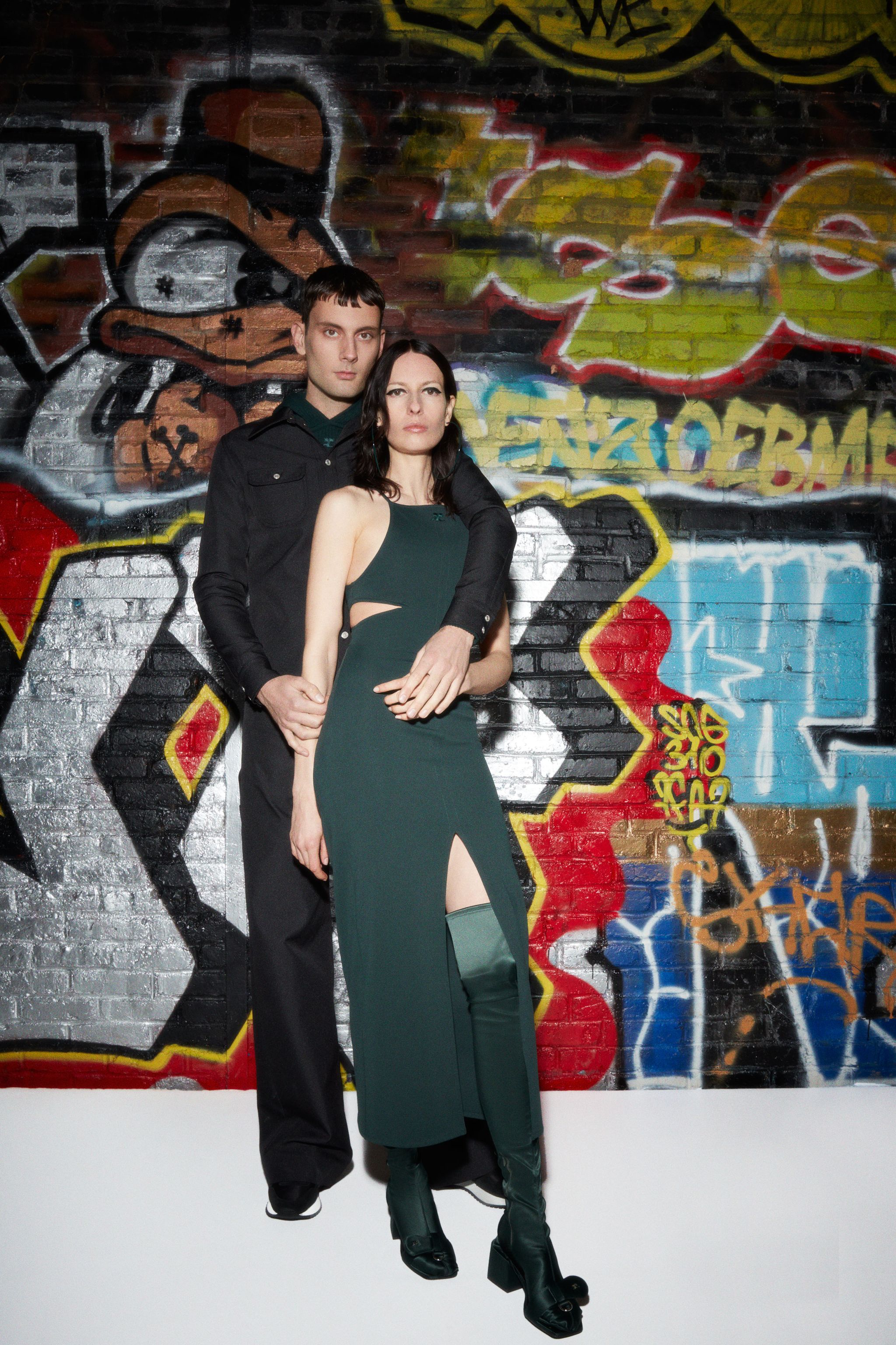 Elegant models in contemporary fashion by Courrèges stand against vibrant graffiti backdrop.