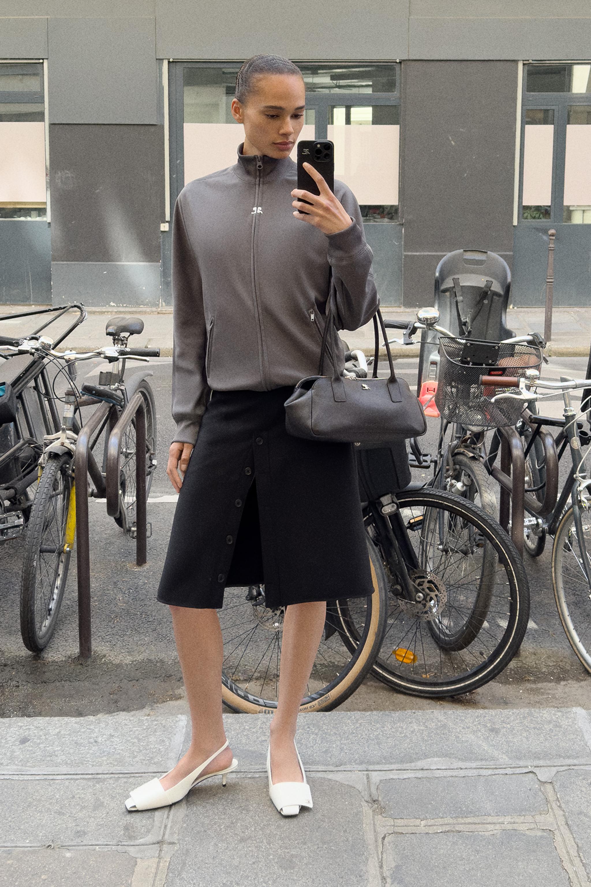Elegant woman in Courrèges fashion, wearing a grey jacket, black skirt, white heels, and carrying a black handbag.