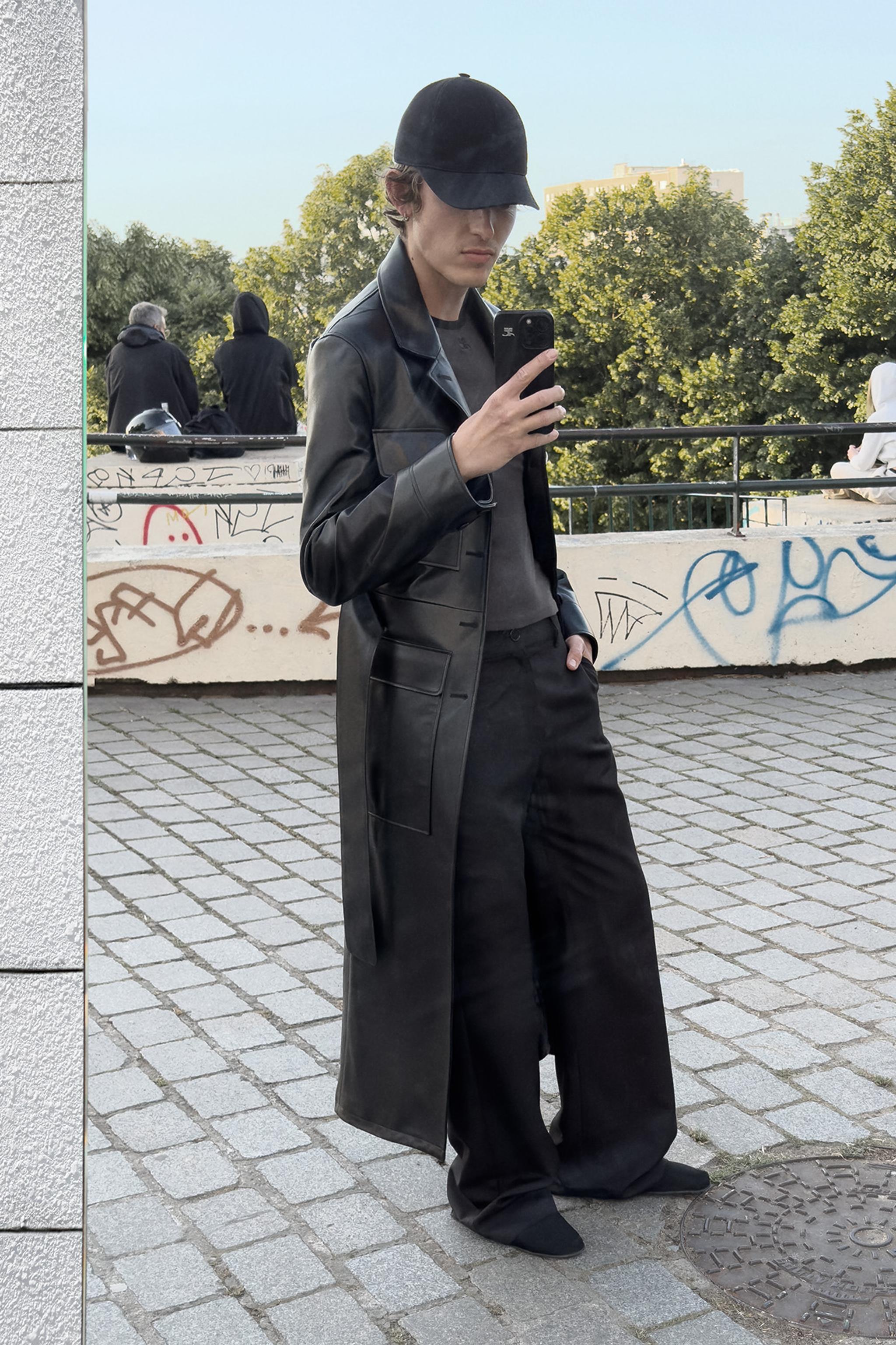 Elegant young man wearing a black leather coat and cap, taking a mirror selfie outdoors.