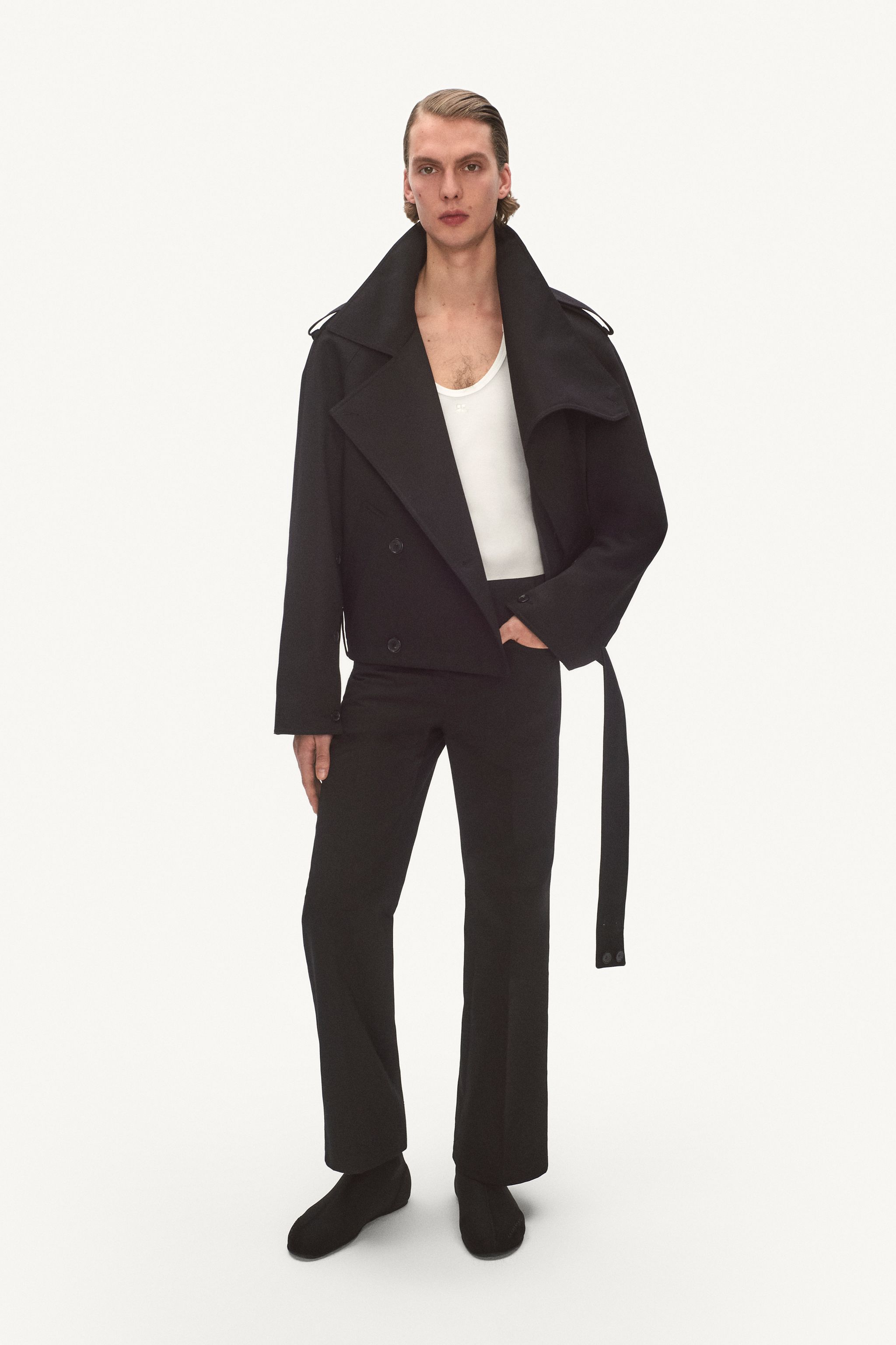 Elegant male model in a black trench coat and tailored trousers, showcasing Courrèges fashion.